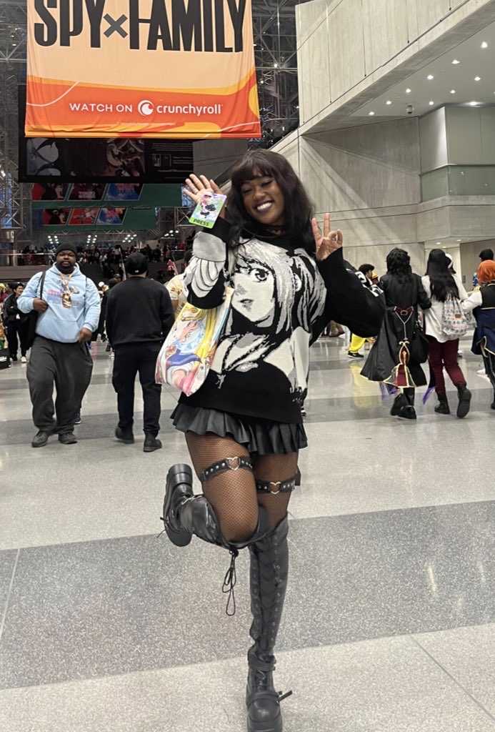 Woman smiling and waving in a busy indoor public space, wearing a graphic sweater, black skirt, and thigh-high boots.