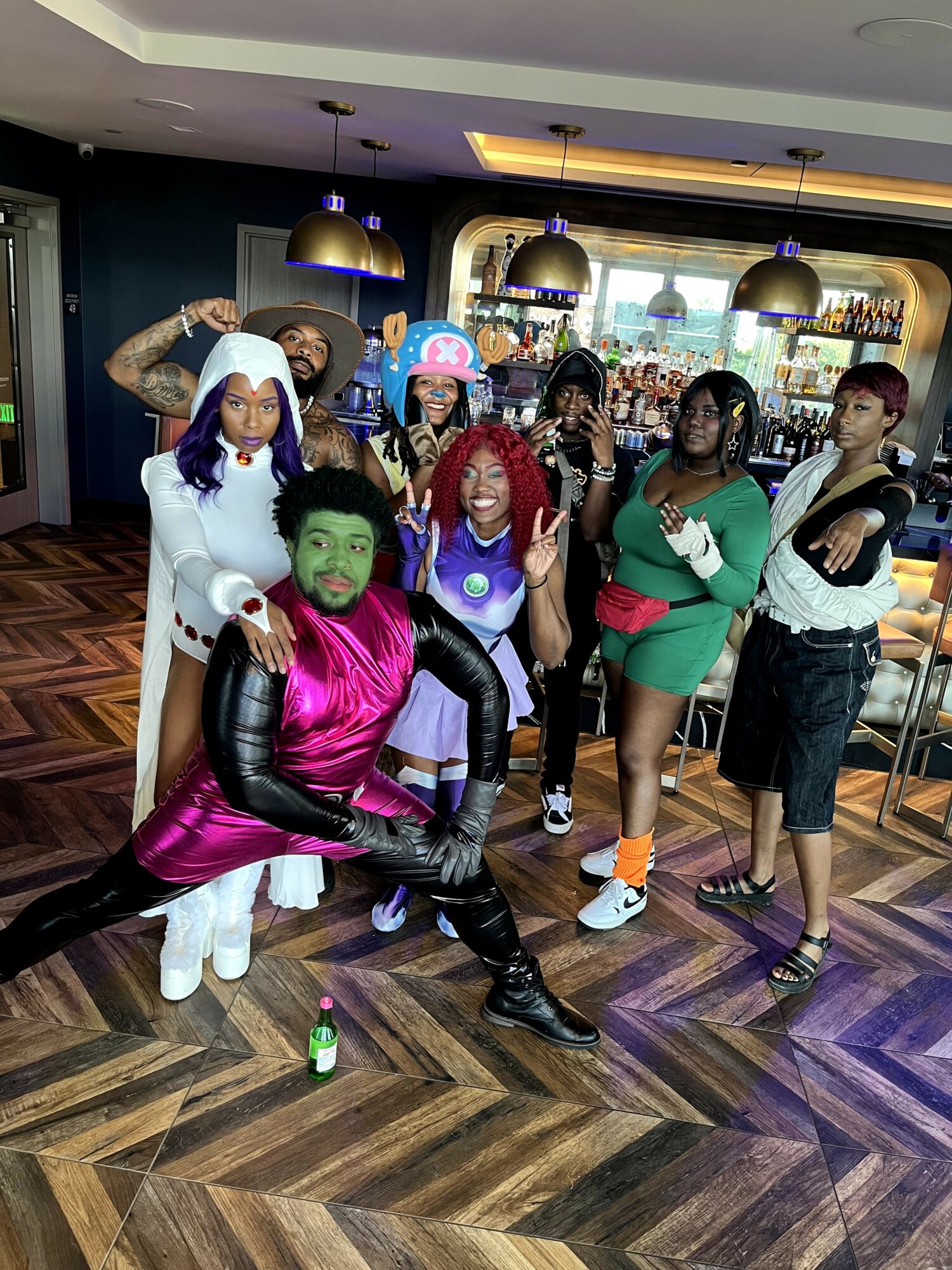 Group of people in colorful costumes and makeup posing indoors with bar and seating area in background.