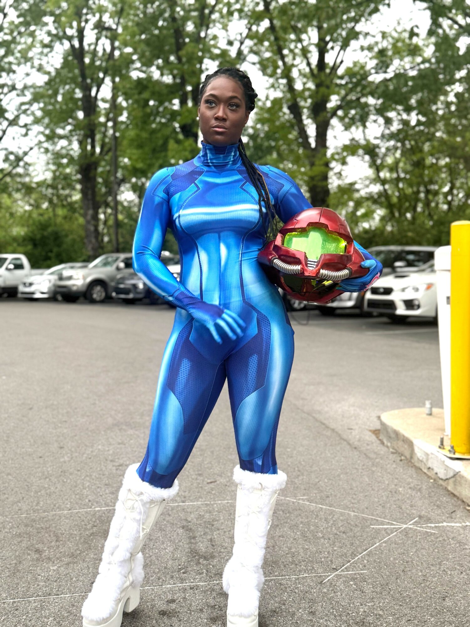 Woman in a shiny blue superhero costume holding a helmet, standing outdoors with trees and parked cars in background.
