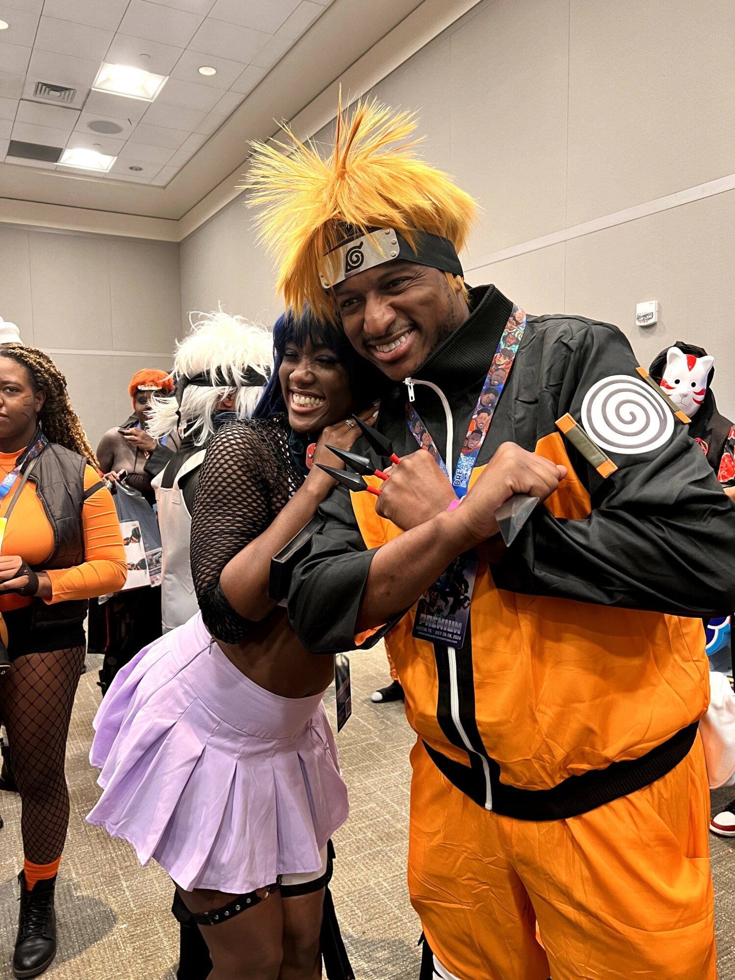 Two people in cosplay costumes smiling and posing together at an indoor event, with other attendees in the background.