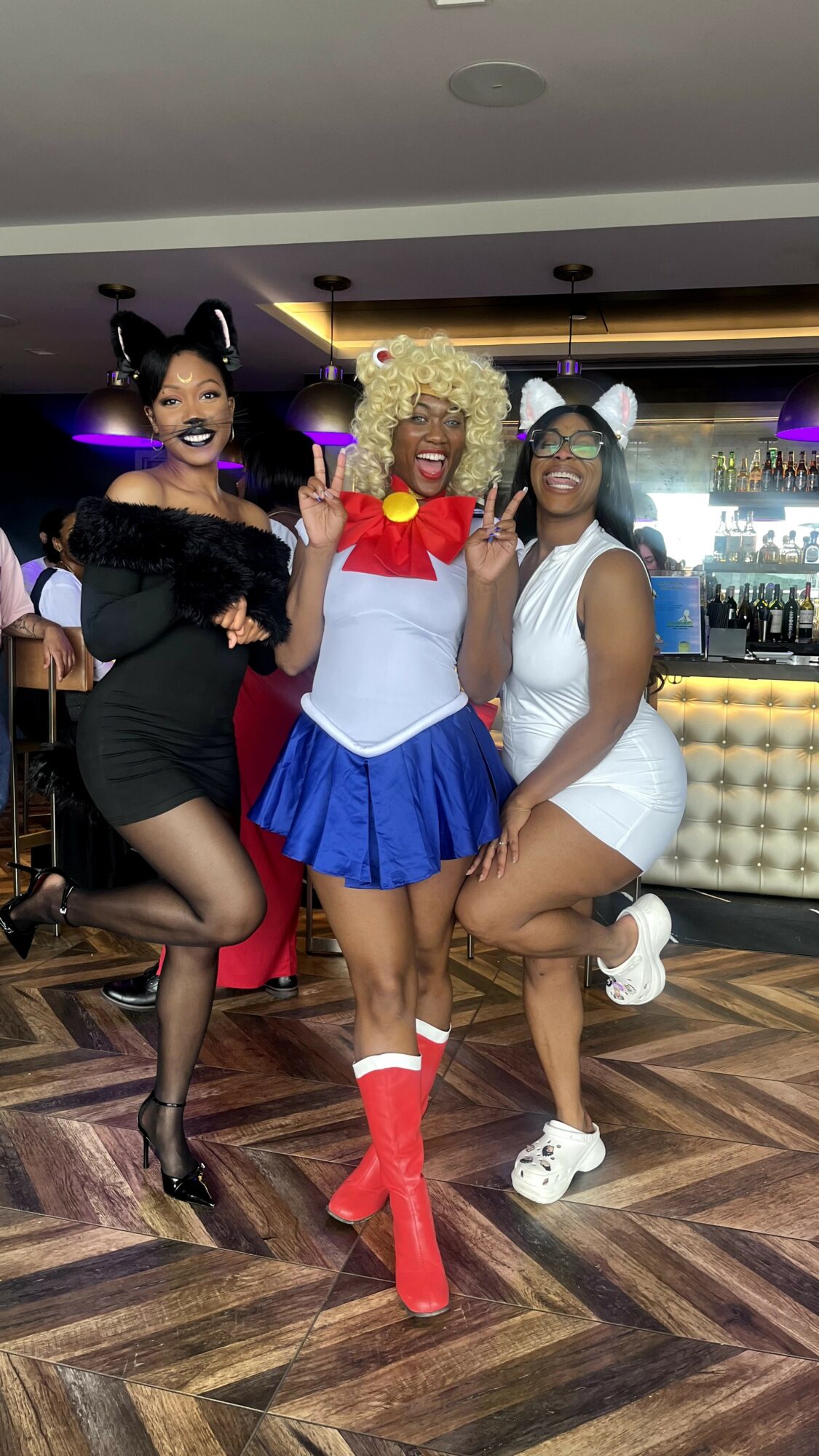 Three women in costumes pose in a bar, smiling, with a bar counter and drinks in the background.