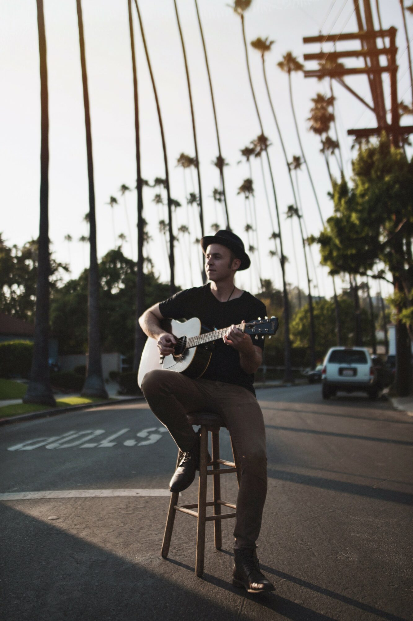 Person sitting on a stool playing guitar outdoors with palm trees and power lines in background.