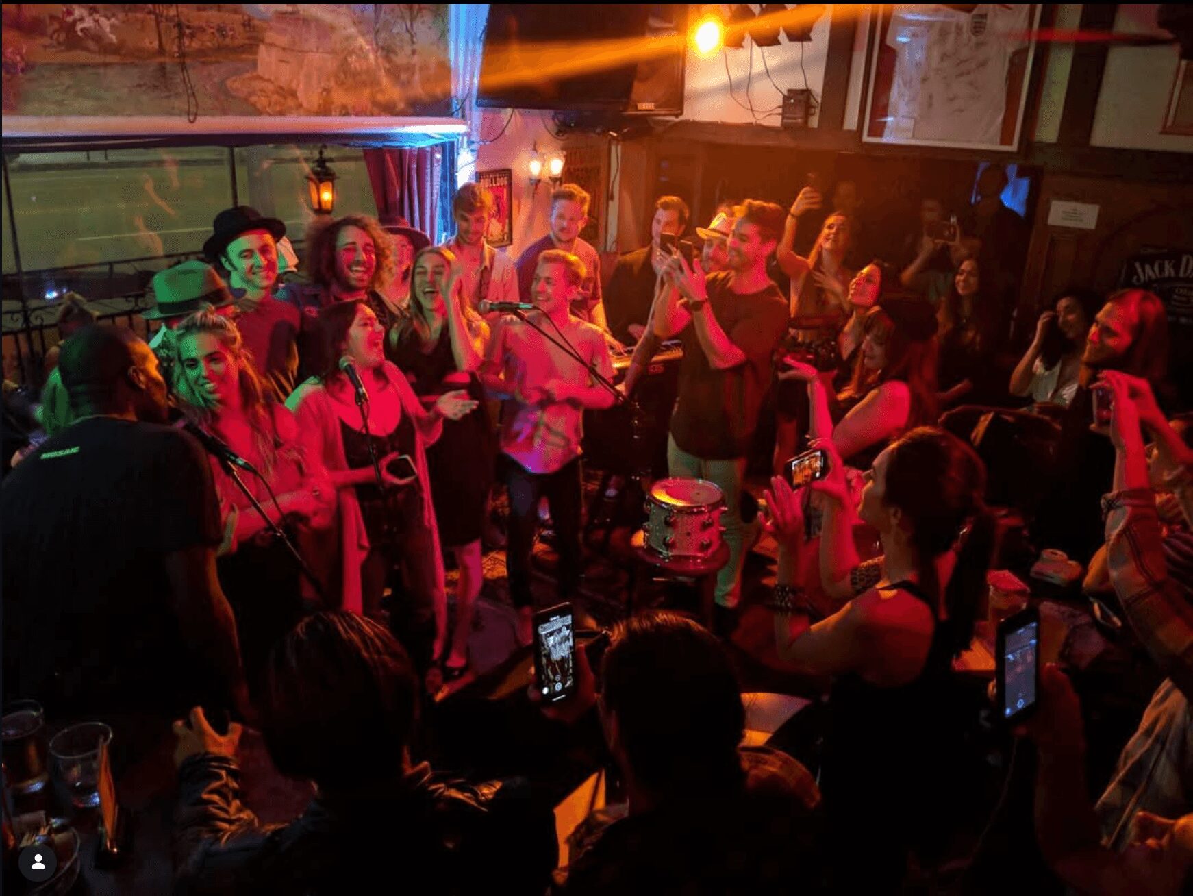 Group of people singing and celebrating around a cake in a lively indoor setting with colorful lighting.