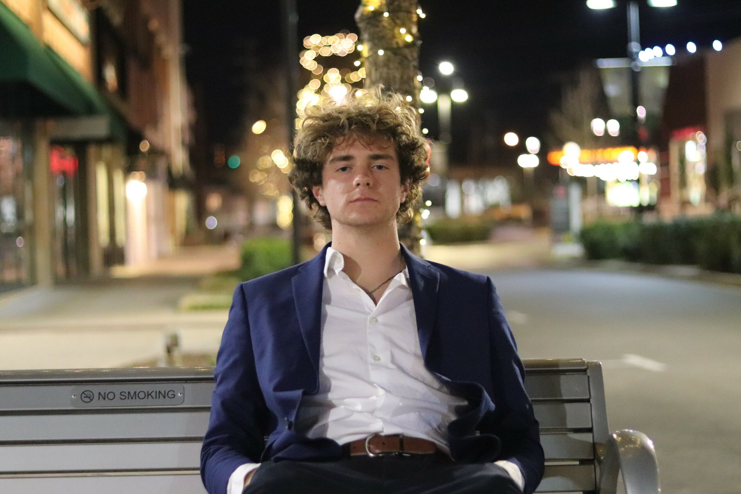 Young man with curly hair in a suit sitting on a bench at night, city street background, lights, and trees.