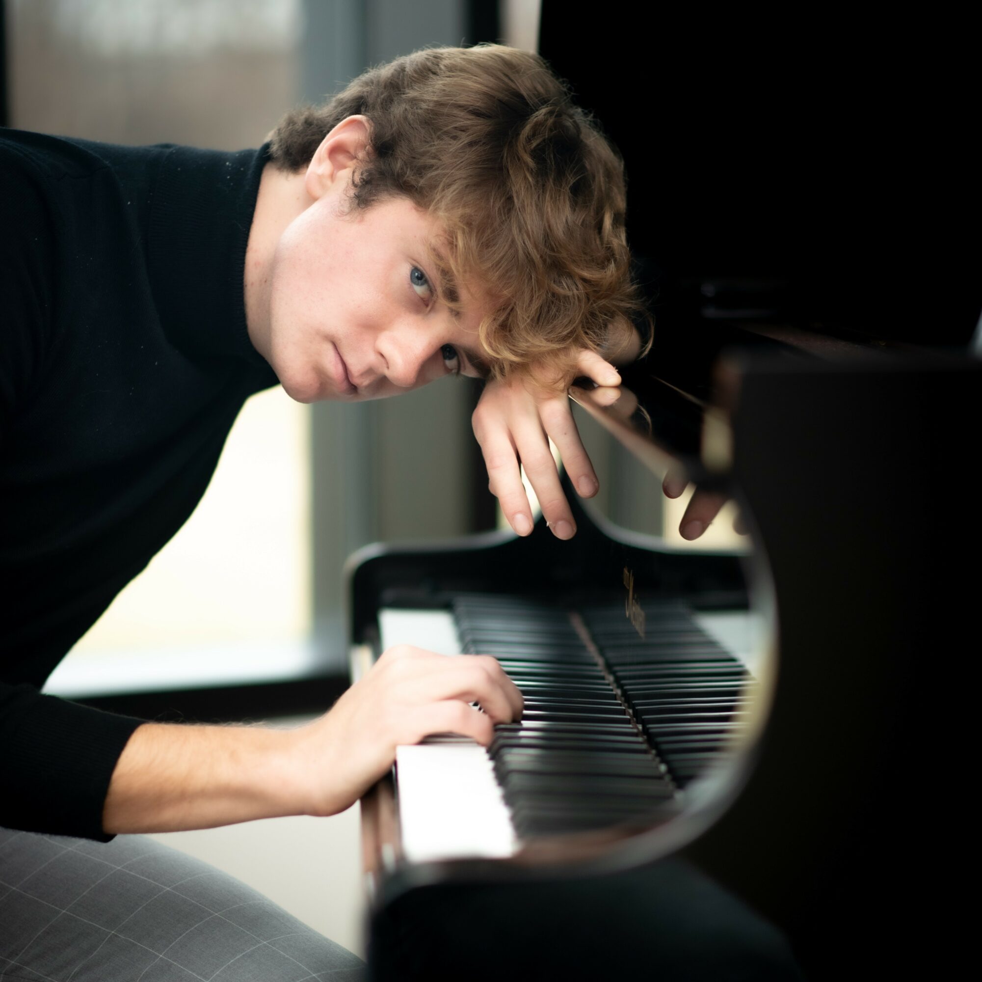 Young man with curly hair leaning on a piano, looking at the camera, in a room with natural light.