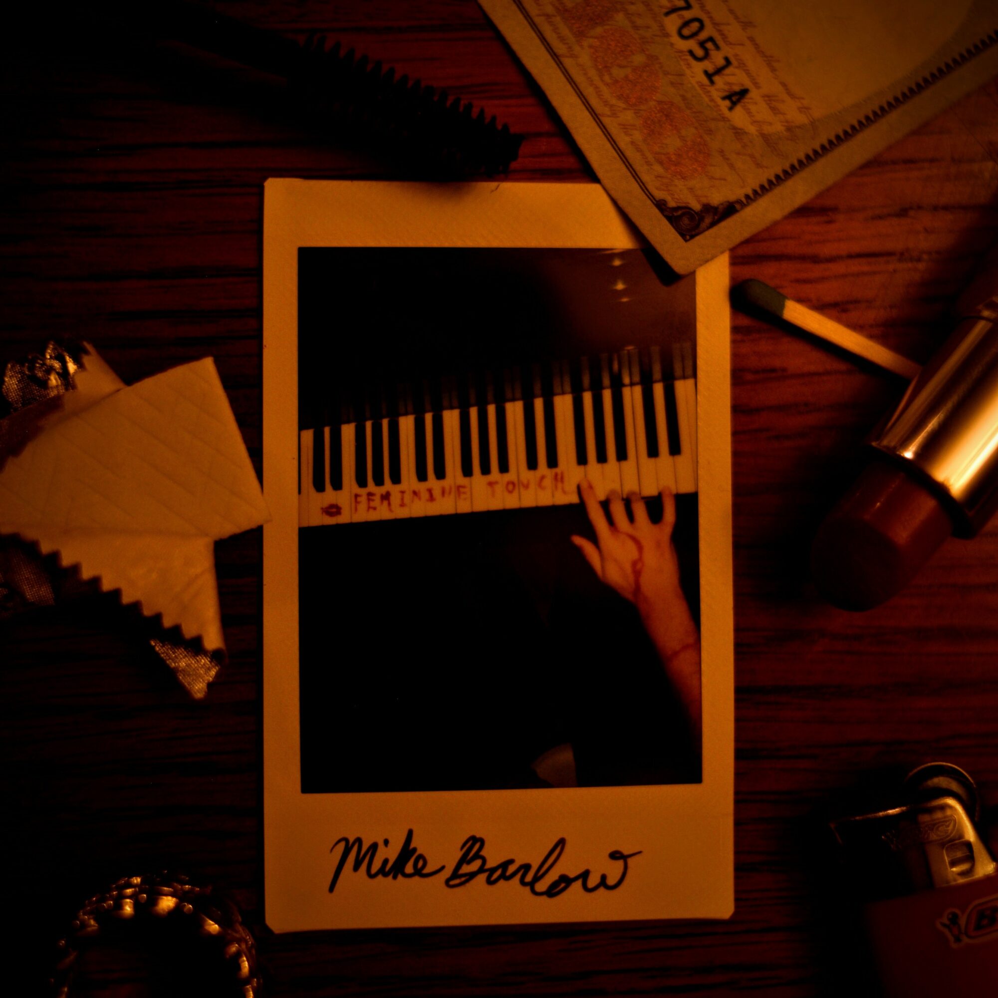Polaroid photo of a person playing piano, signed 'Mike Barlow', on a wooden surface with various objects around.