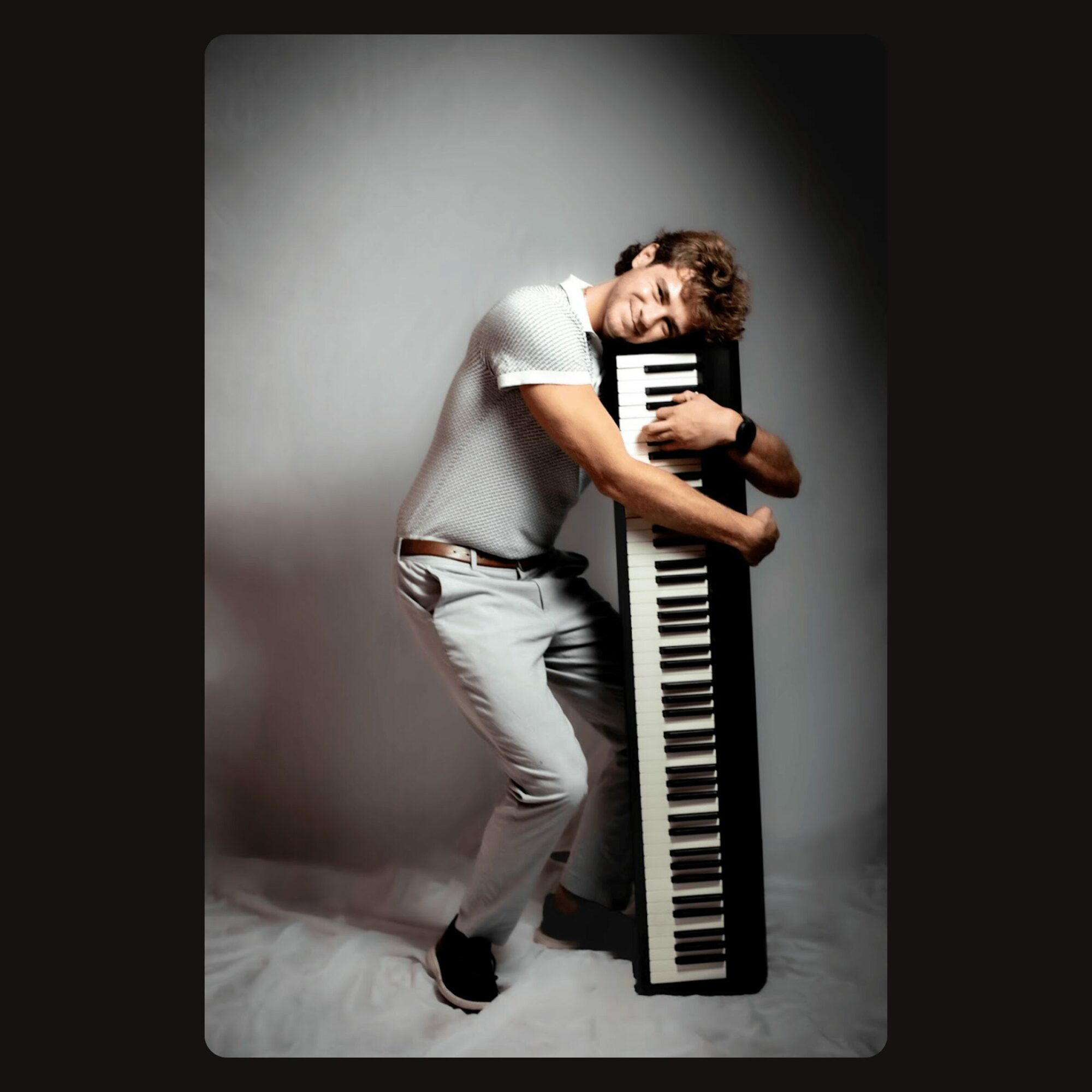 Young man hugging a large keyboard, smiling, in a studio with a gray background.