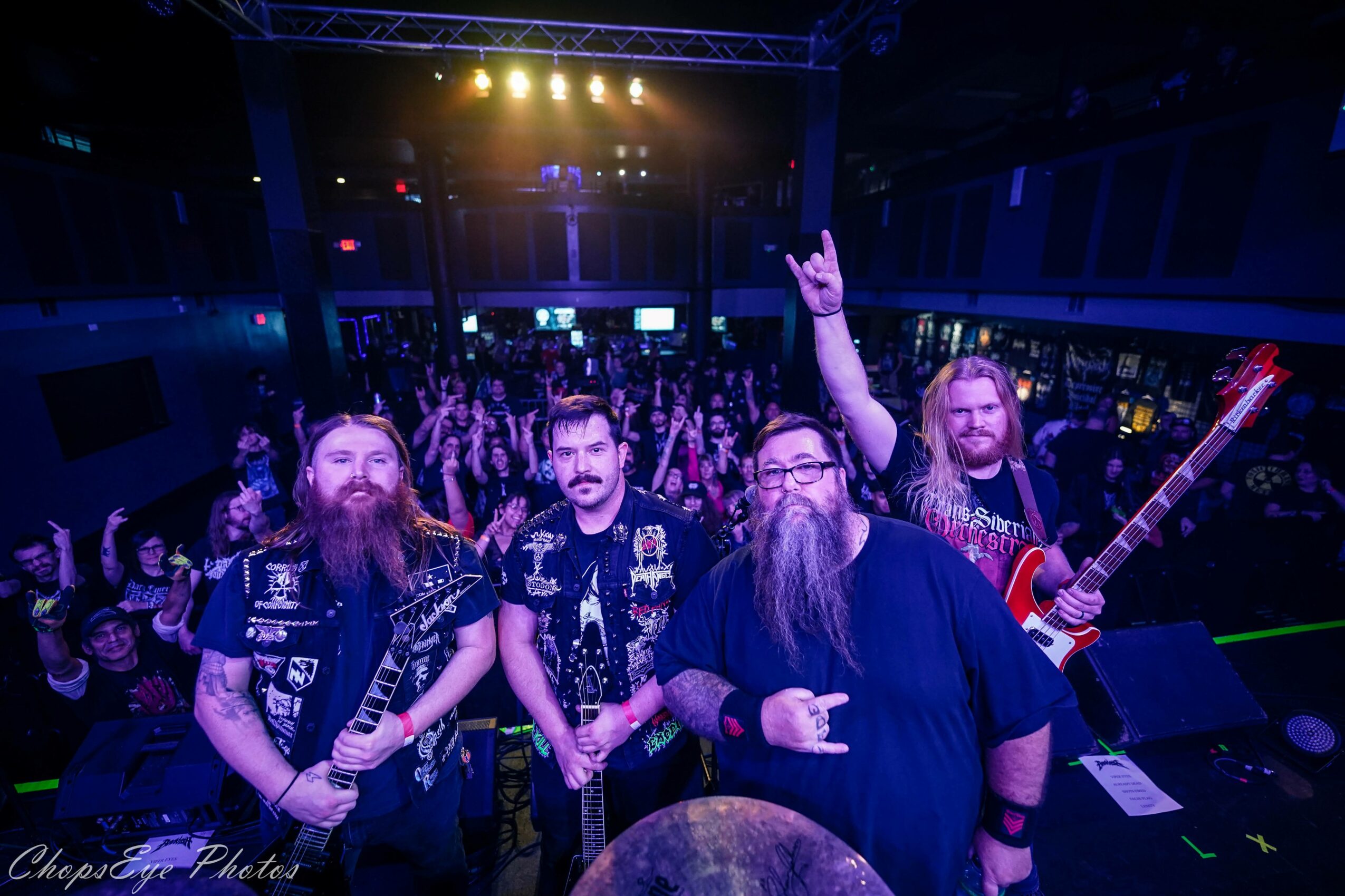 Four band members on stage with a crowd in the background, stage lights shining down, one person raising a hand.