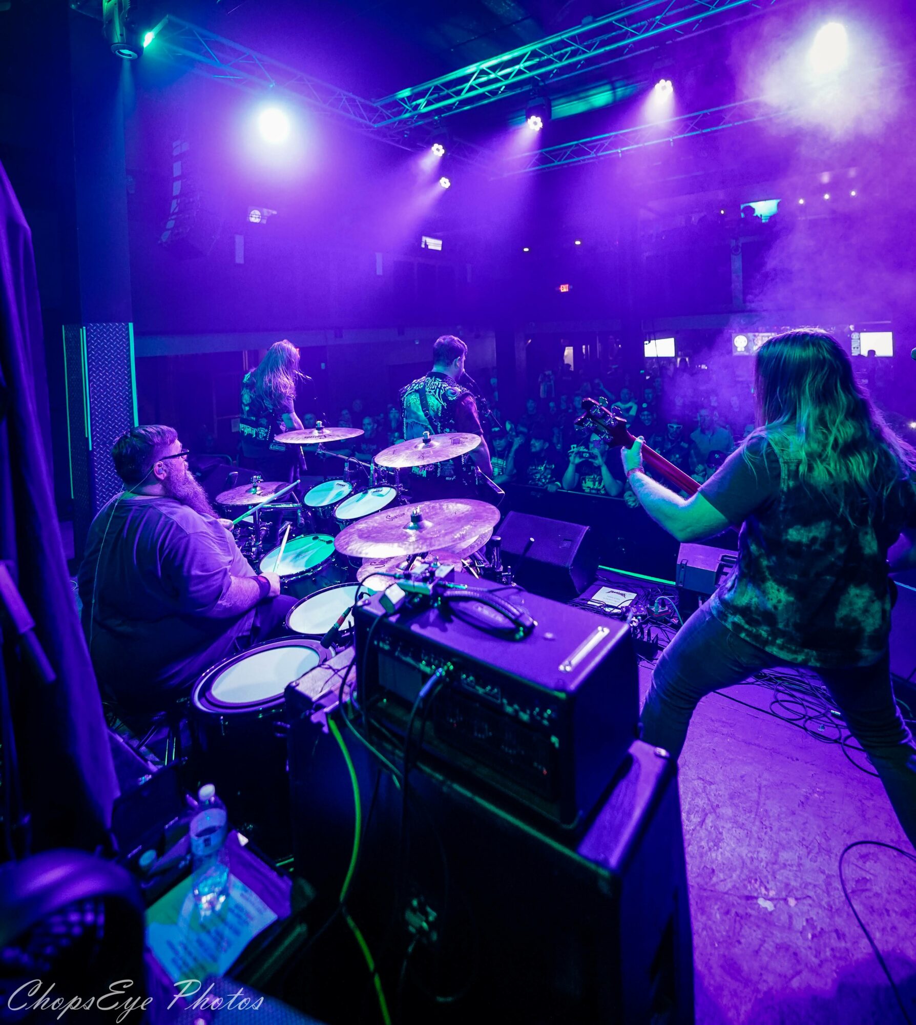 Band performing on stage with drums and a singer, colorful stage lights, audience in background, in a concert venue.