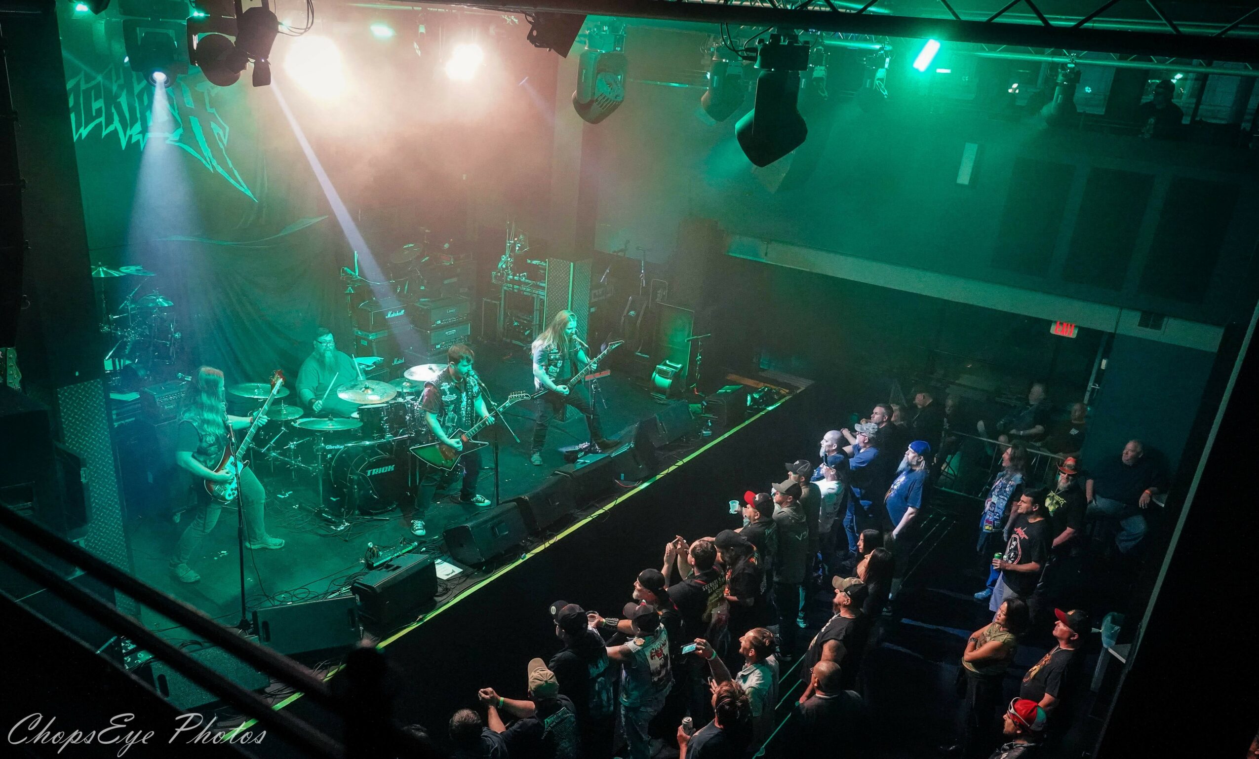 Band performing on stage with audience watching in a dimly lit venue, green and purple lighting effects, audience standing and seated.