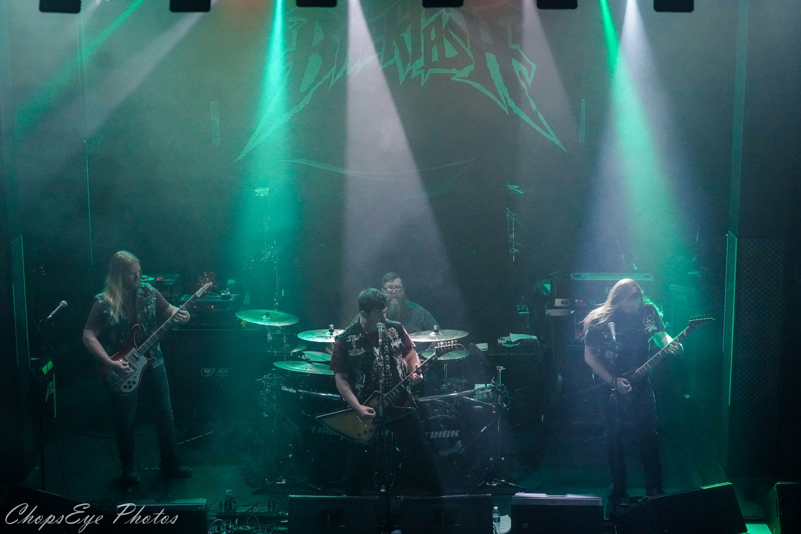 Four musicians perform on stage with colorful lighting and a dark background.