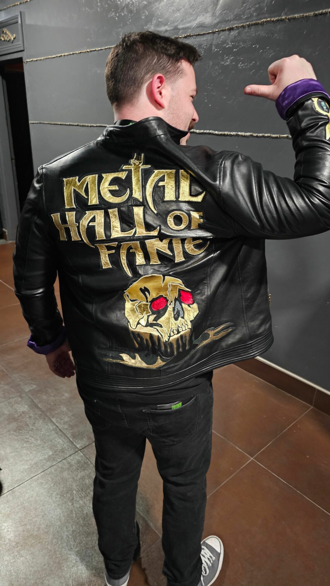 Person standing sideways, wearing a black jacket with gold and red skull design, black pants, and sneakers, indoors.