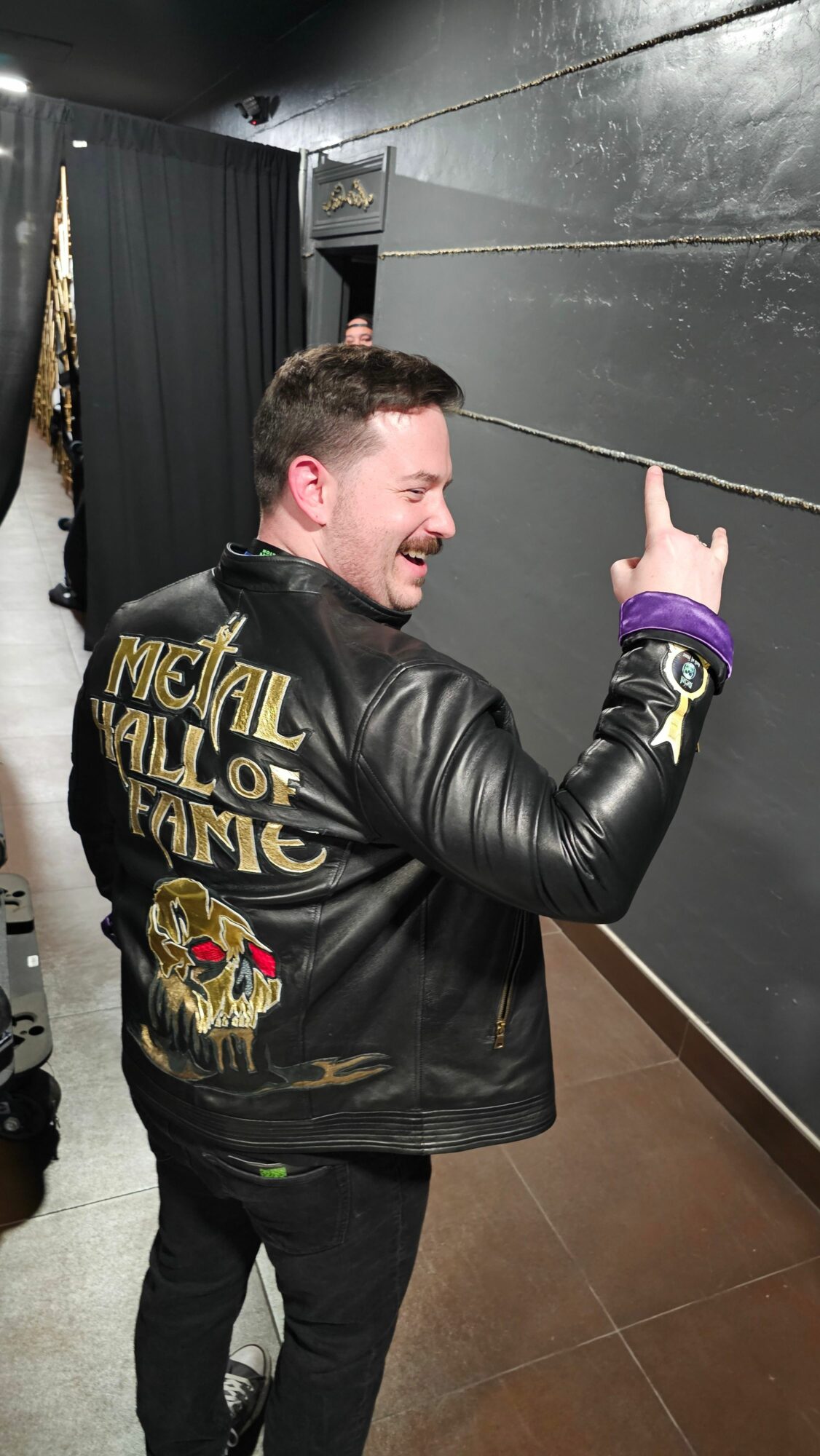 Person with short hair wearing a black leather jacket with patches, pointing upward, standing indoors against a dark wall.