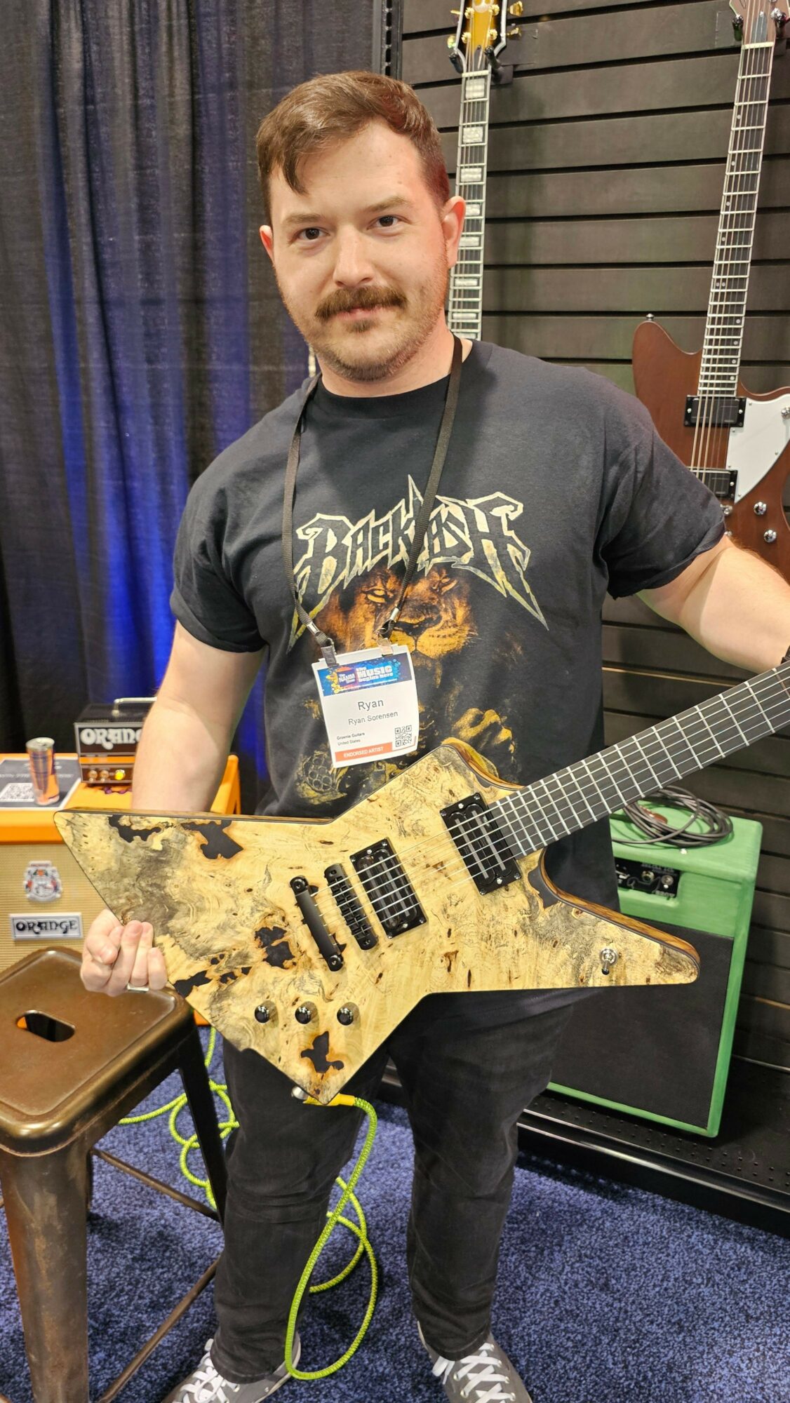 Young person holding a guitar with a distressed finish, wearing a graphic t-shirt, standing in a room with musical equipment.