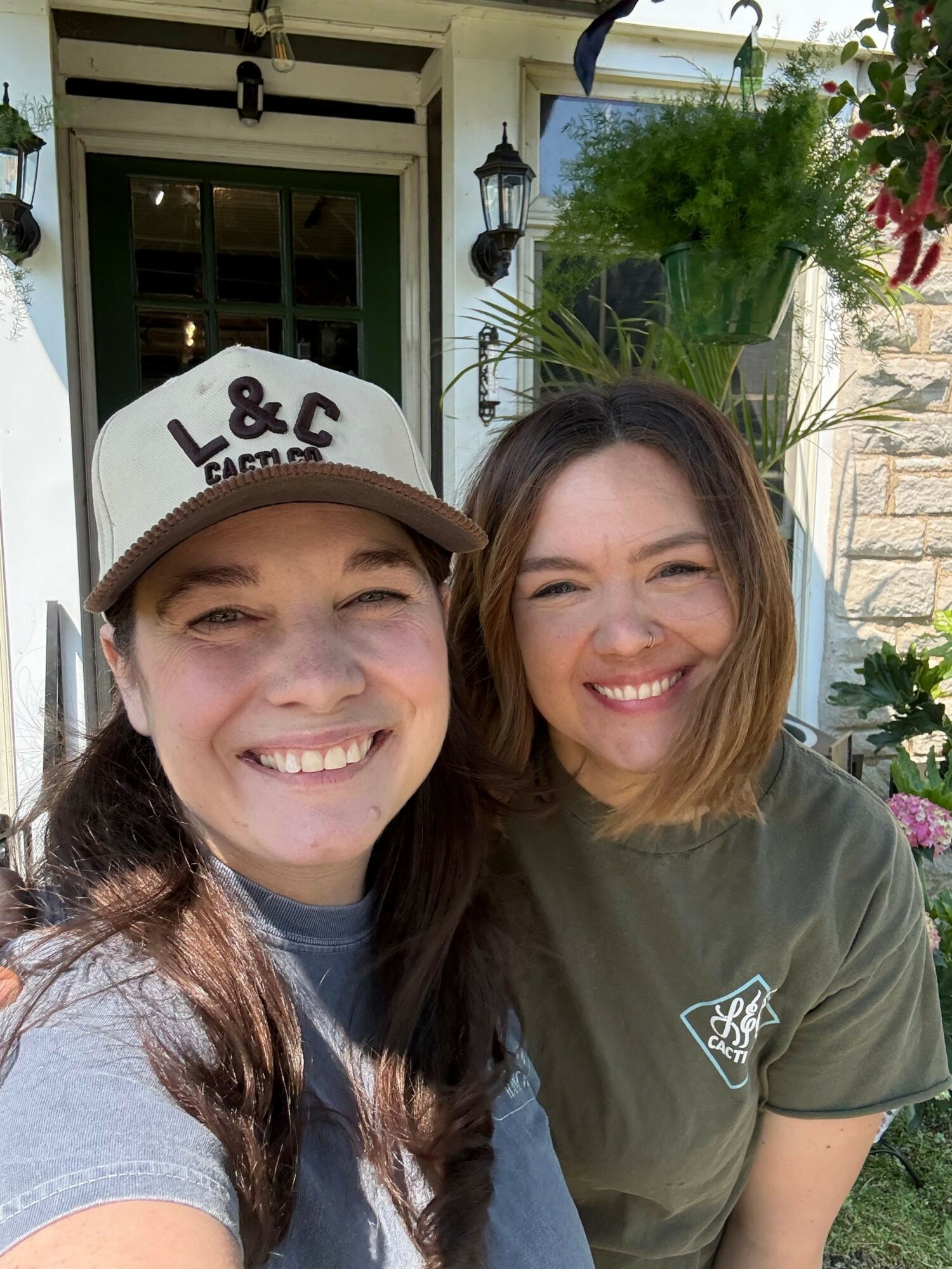 Two smiling women outdoors, one wearing a baseball cap and the other with long hair, in front of a house with plants.