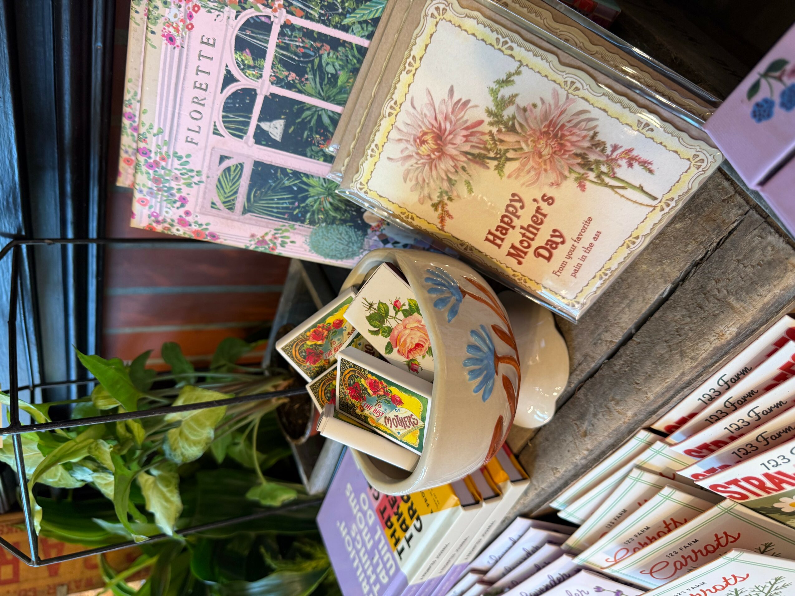 Decorative items including a floral greeting card, a mug with colorful designs, and a small book or card, with green plant leaves nearby.