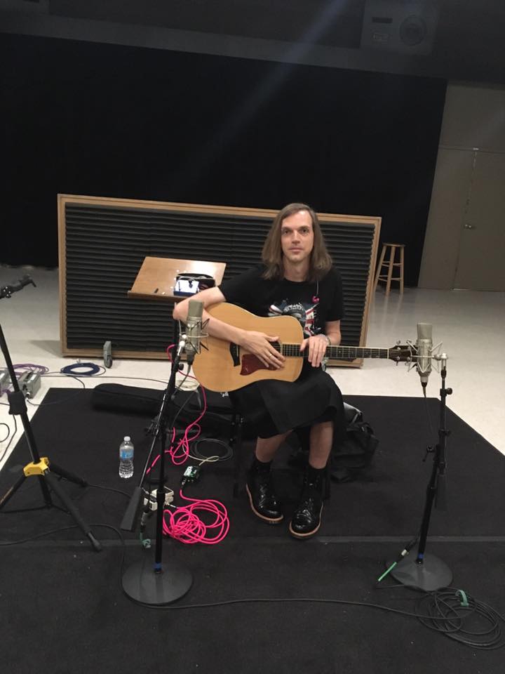 Person with long hair sitting on a stool, holding an acoustic guitar, in a music studio with microphones and equipment.