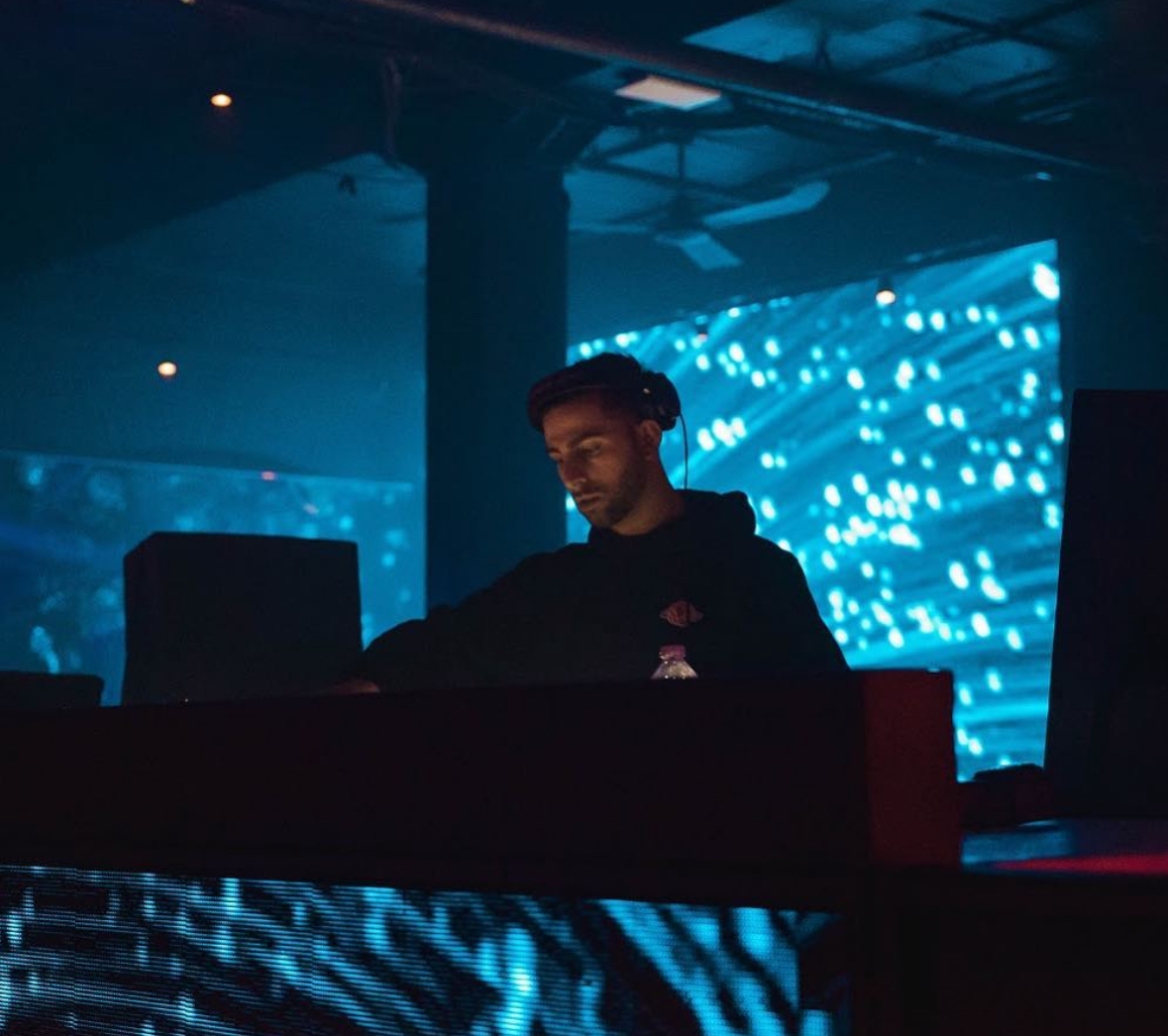 Person with glasses working at a DJ booth in a dark, illuminated venue with blue digital background.