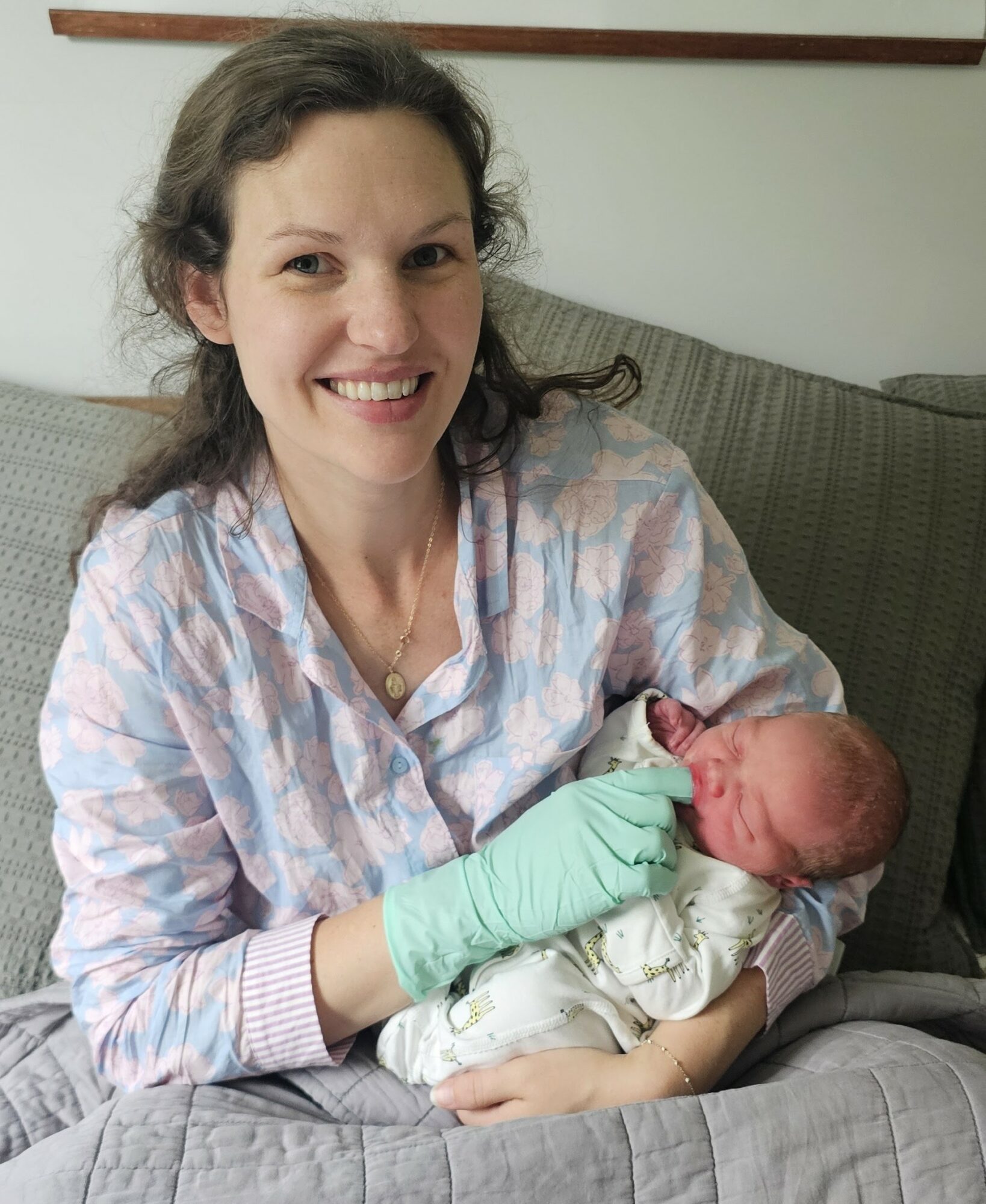 Woman holding a newborn baby, smiling, sitting on a couch in a room with a light-colored wall.