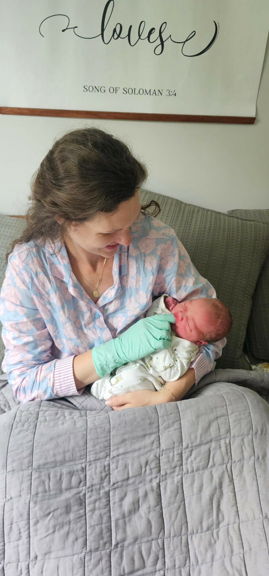 Woman in hospital gown holding a newborn baby, sitting on a bed with a gray quilt, in a room with wall art.