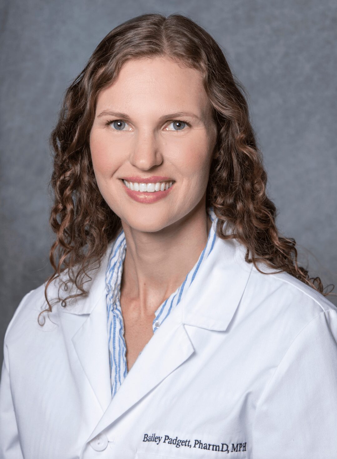 Portrait of a woman with curly hair wearing a white lab coat, smiling, against a gray background.
