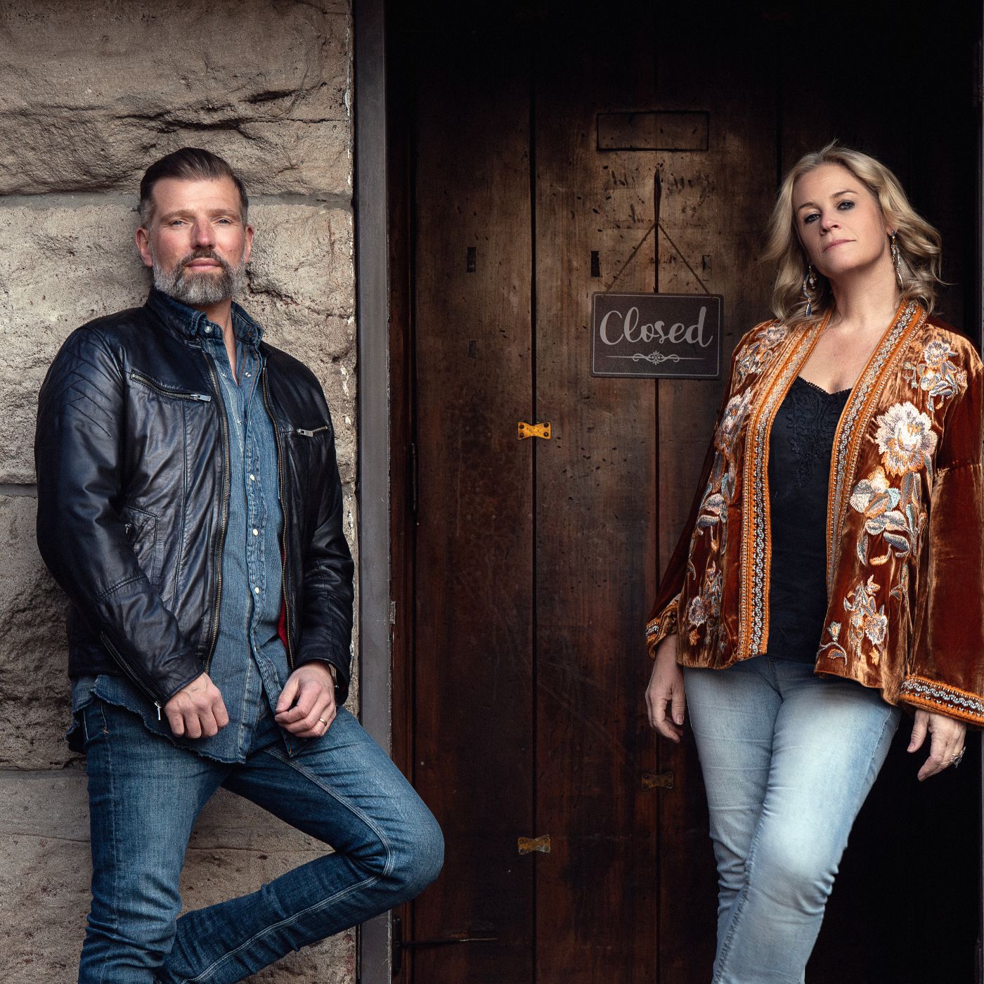 A man and woman stand outside a door with a closed sign, posing confidently. The man wears a leather jacket, jeans, and has a beard. The woman wears a patterned jacket and light jeans.