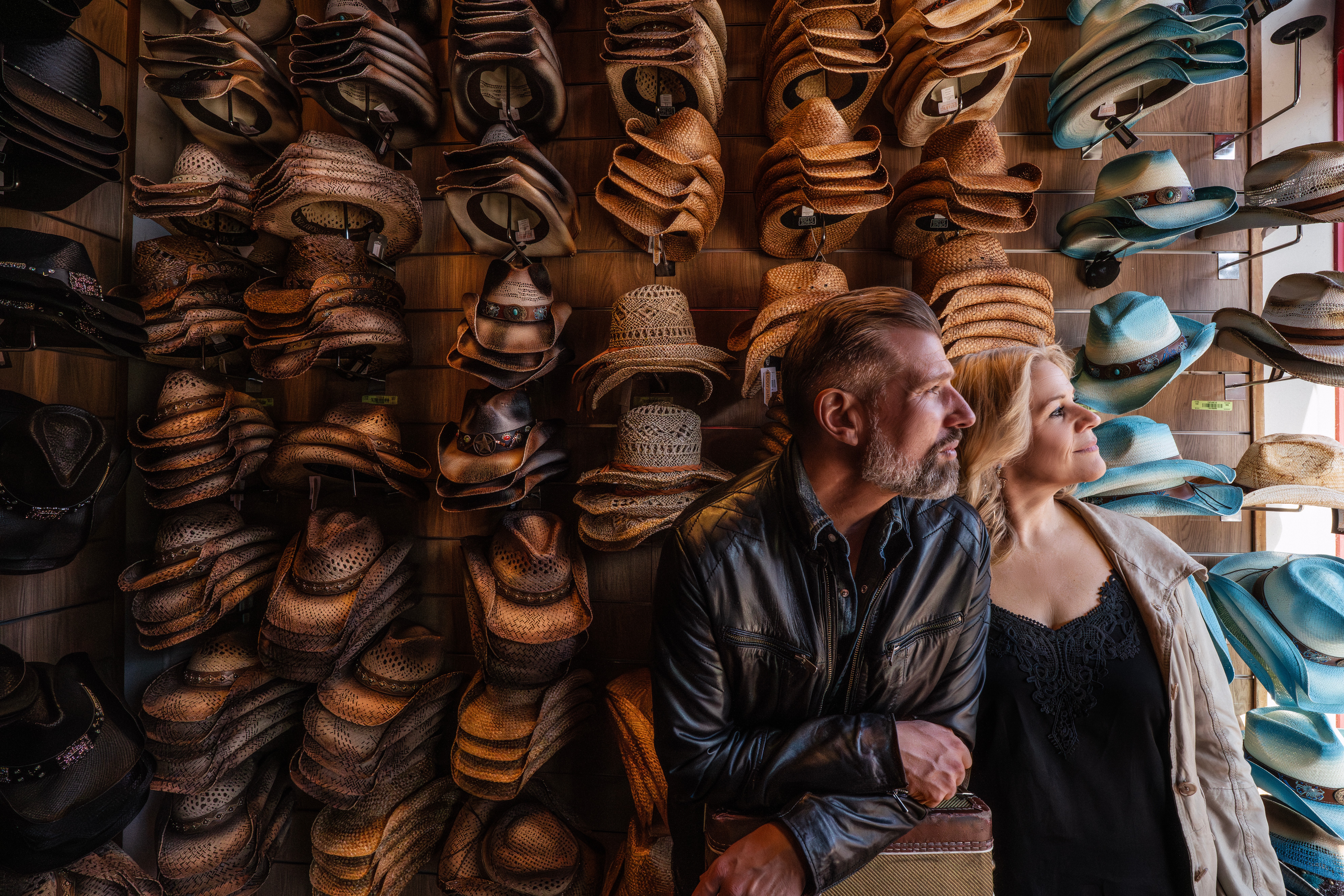 Two people sit in front of a wall display of various hats, looking to the right, with hats stacked behind them.
