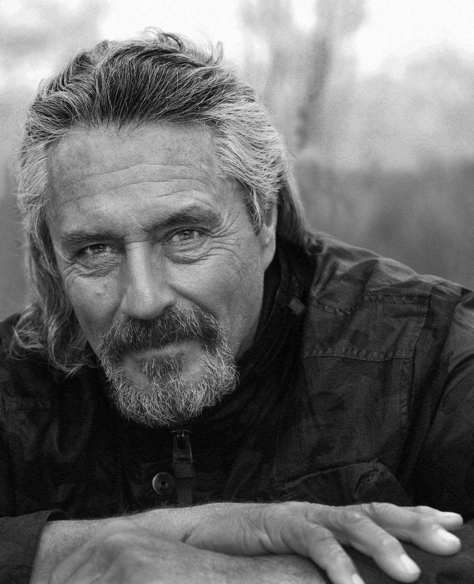A black and white portrait of a middle-aged man with long hair and a beard, outdoors with blurred background.