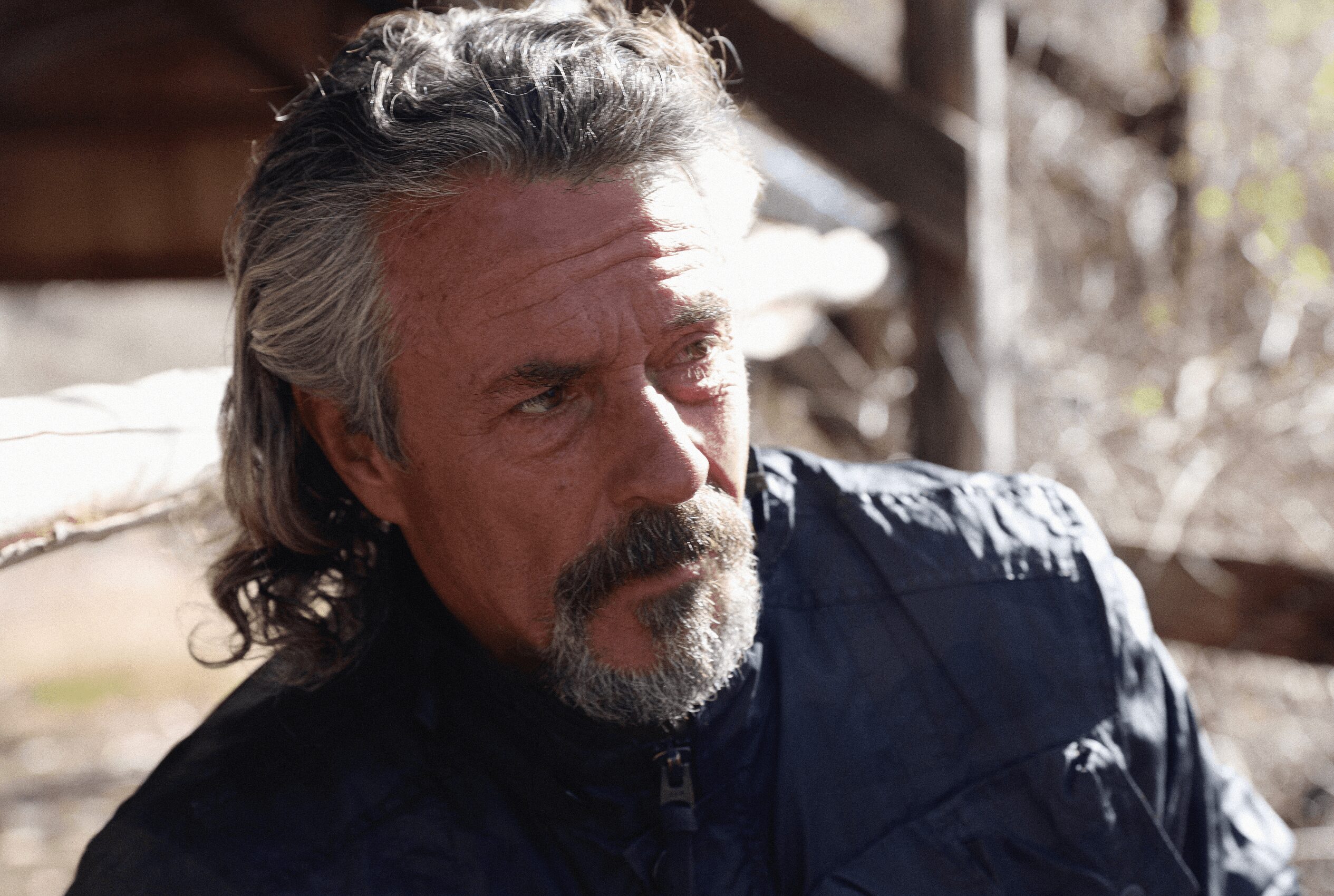 Man with gray hair and beard outdoors, wearing a dark jacket, looking to the side.