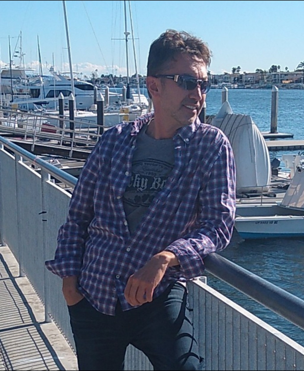 Man wearing sunglasses and a checkered shirt leaning on a marina railing with boats in the background.