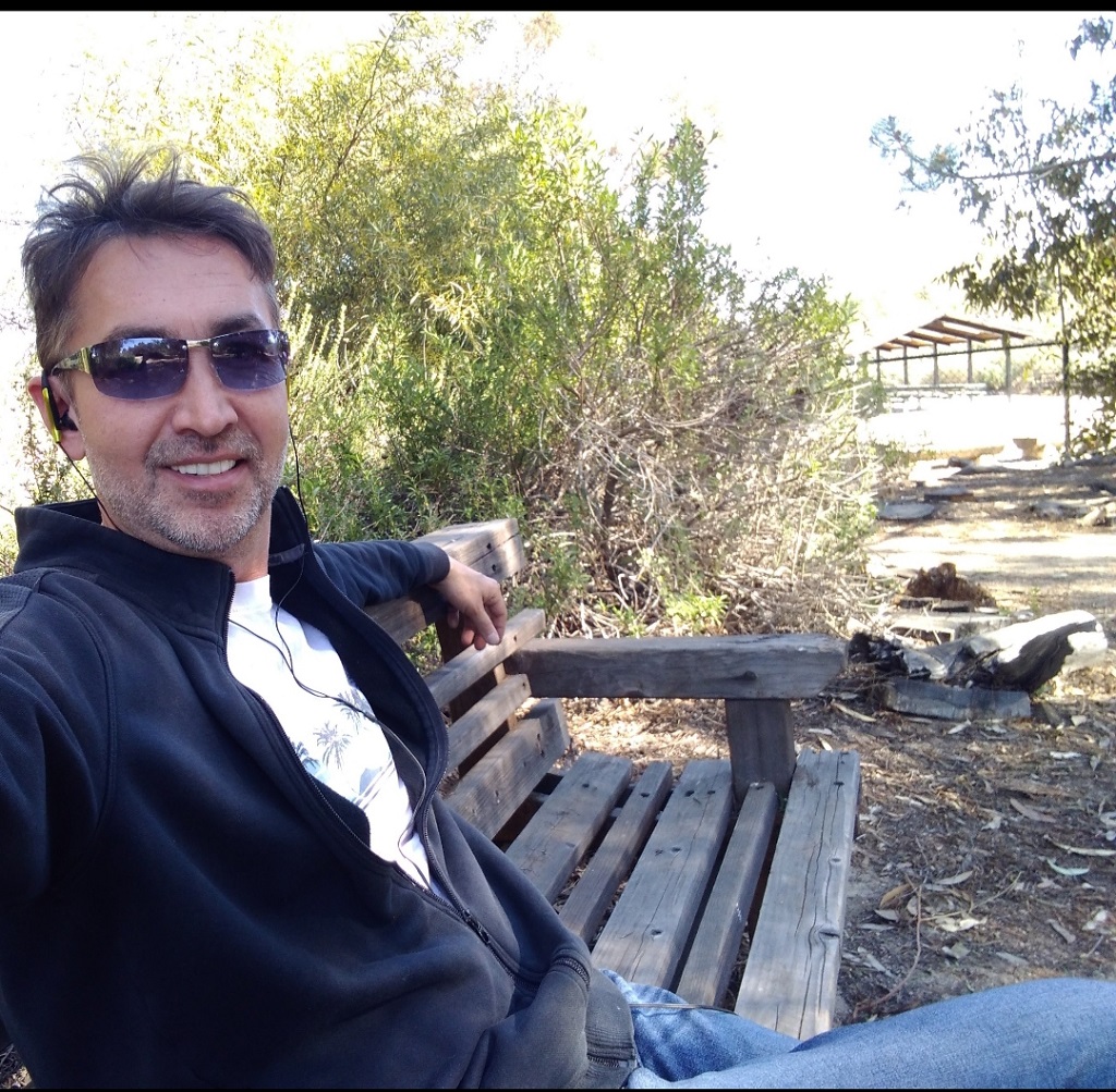 Man sitting on a park bench outdoors, wearing sunglasses and a black jacket, smiling at the camera.