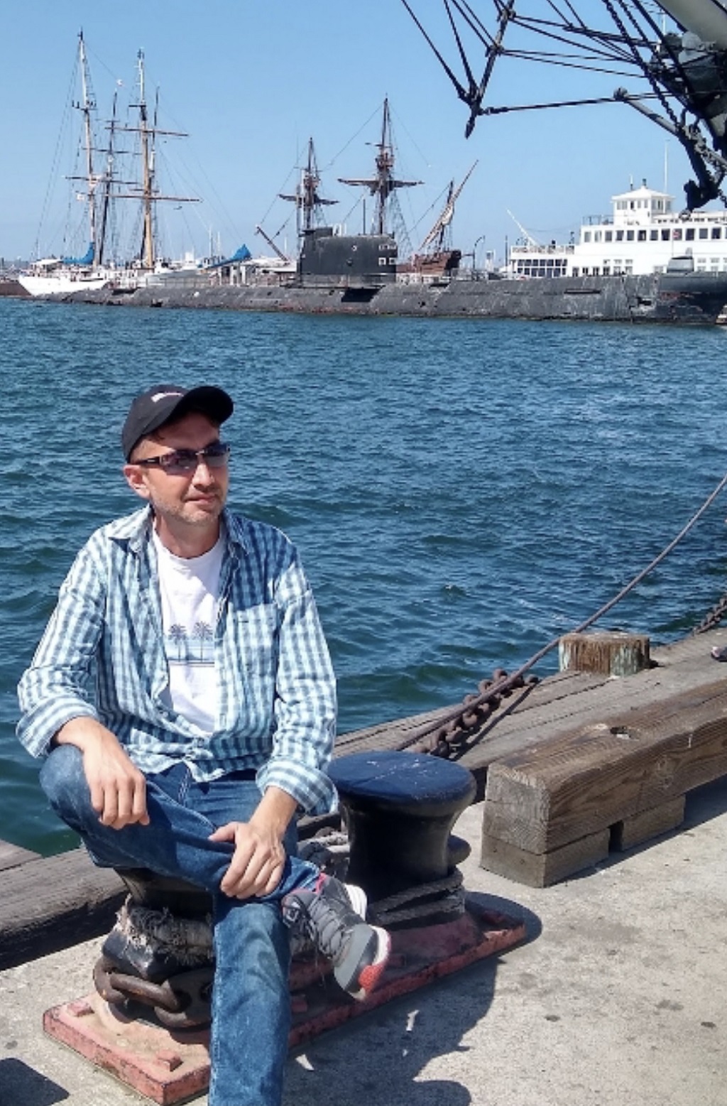 Person sitting on a dock near water with ships in the background, wearing a cap and plaid shirt.
