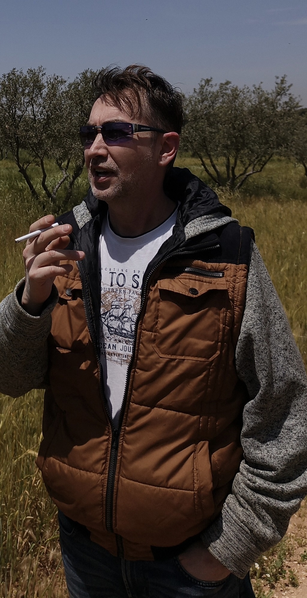 Person wearing sunglasses and a brown and gray jacket, standing outdoors in a grassy area with trees.