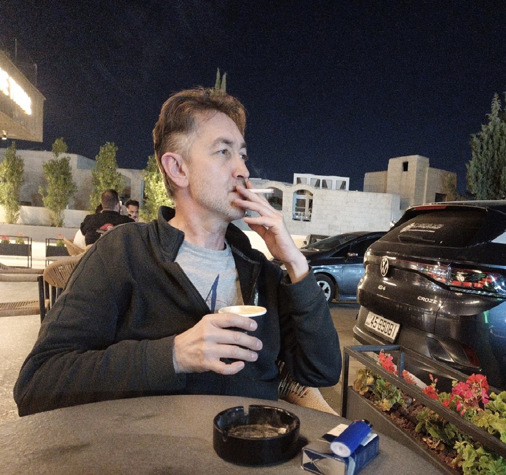 Man sitting outdoors at night, smoking and holding a cup, with cars and buildings in the background.