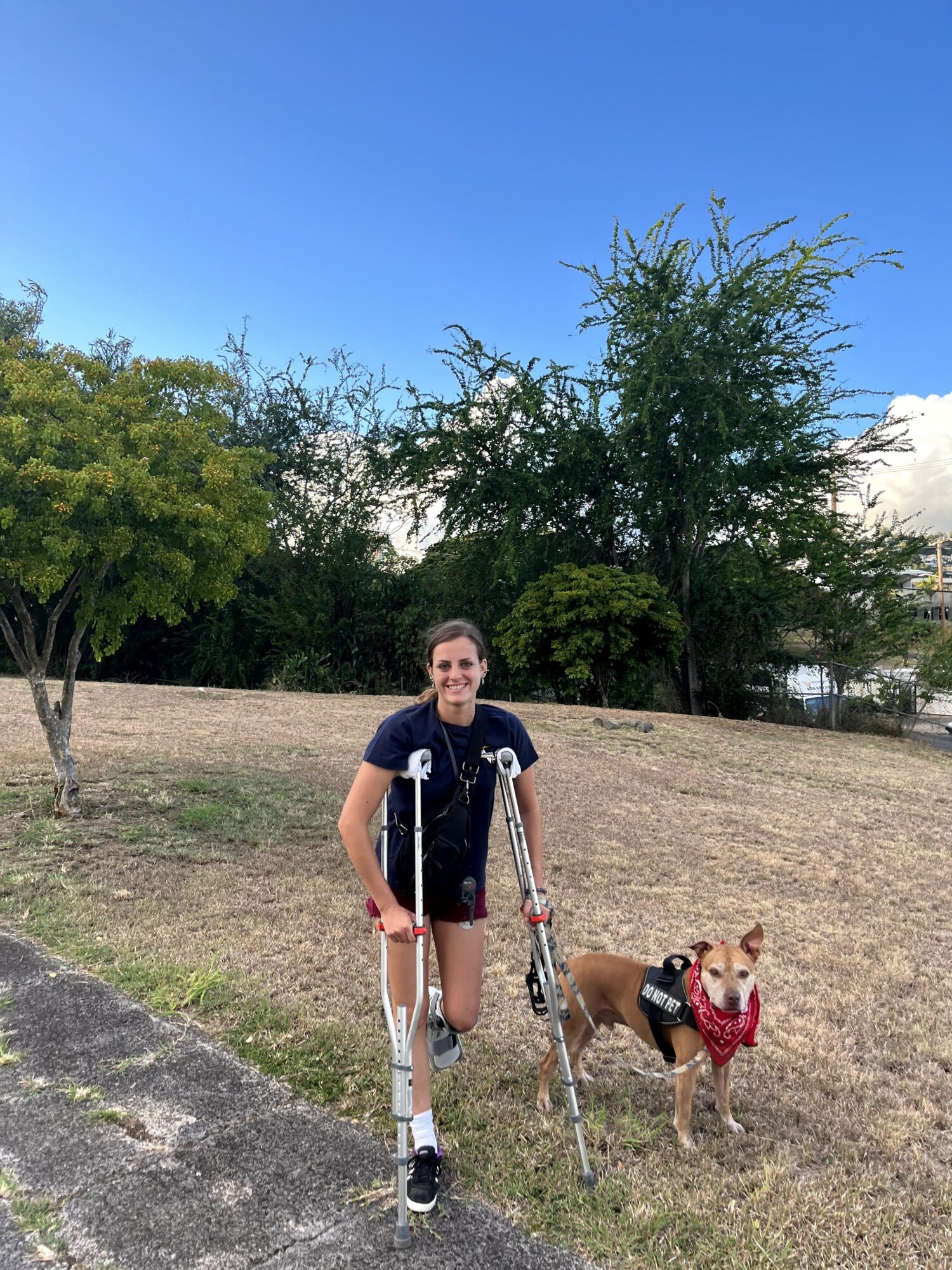 Person smiling with crutches standing on grass with dog wearing a red harness, trees and blue sky in background.