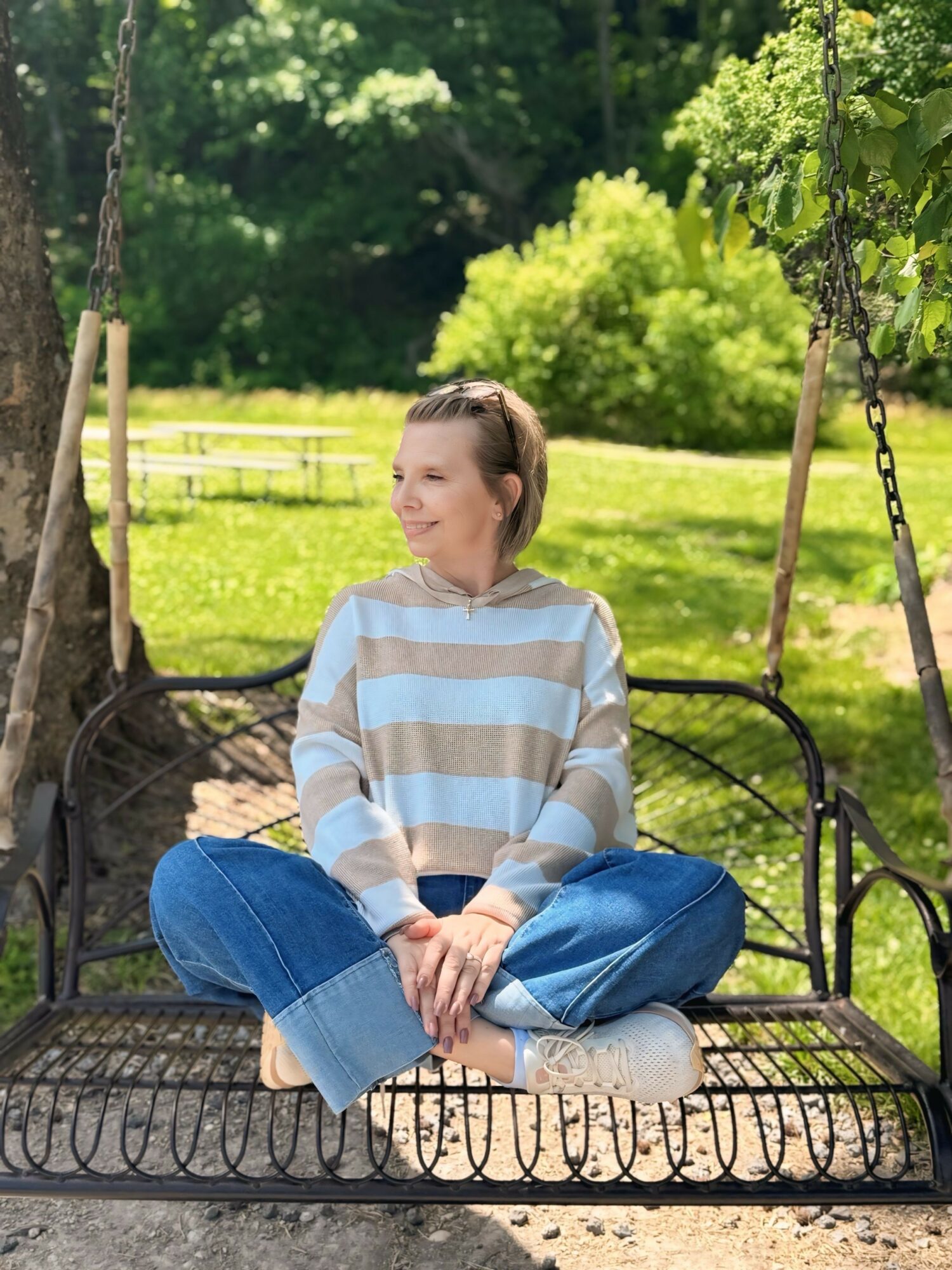 Woman sitting on a swing outdoors, smiling, with trees and a grassy field in the background.