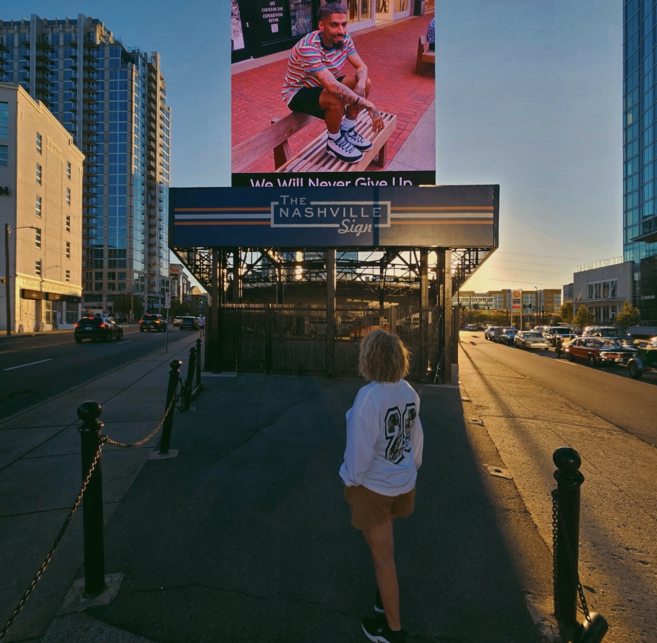 Person walking towards a Nashville sign with tall buildings on both sides during sunset.