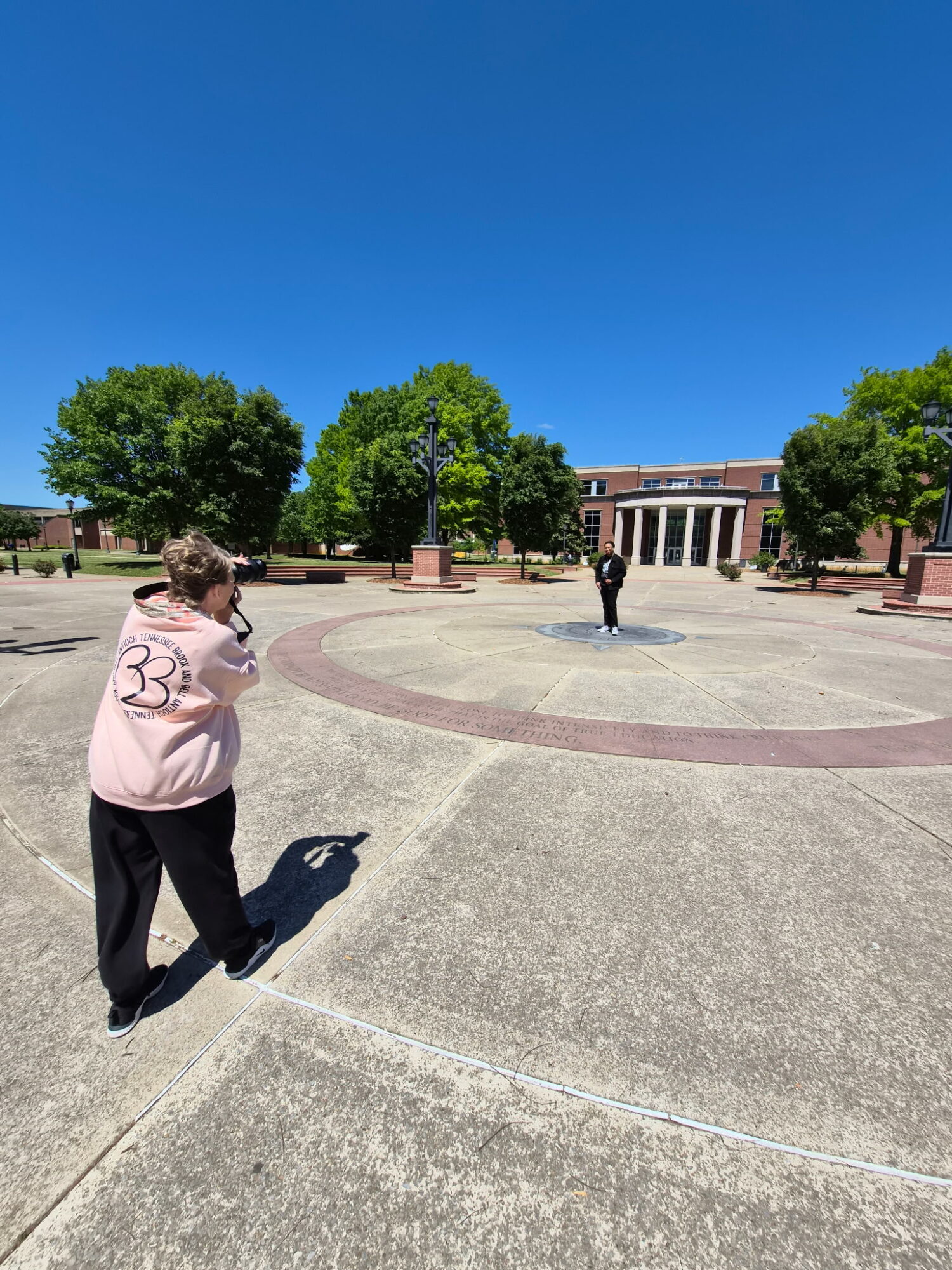 Person standing outdoors on a paved area with trees and a building in the background, under a clear blue sky.