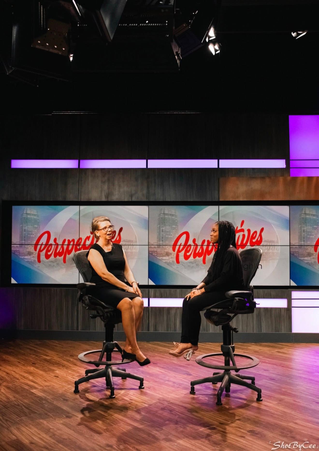 Two women sit on chairs facing each other in a studio with a Perspex logo background, wooden floor, and studio lights.