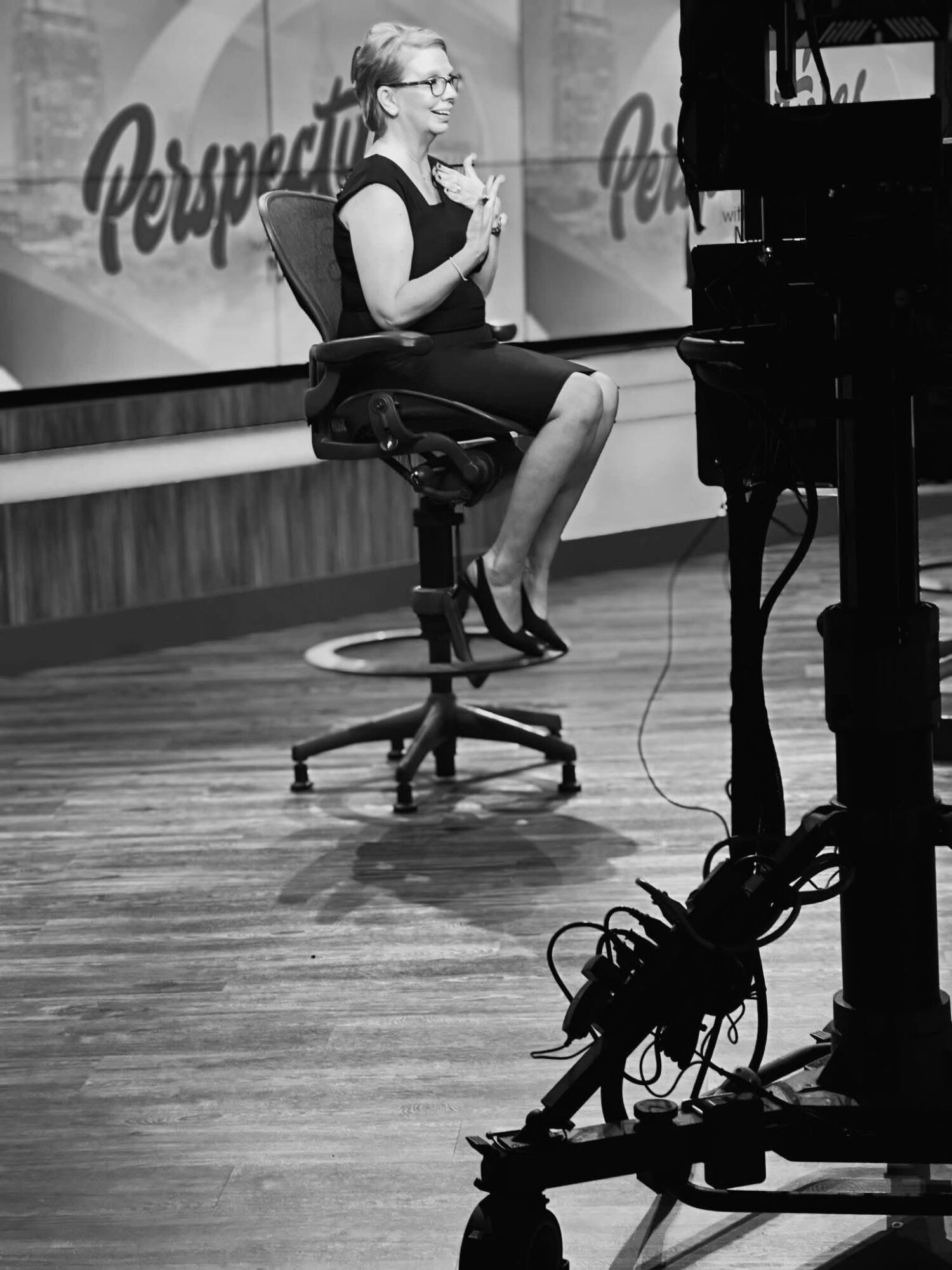 Woman sitting on a stool, holding a microphone, near a piano on a stage with a backdrop reading 'Perspective'.