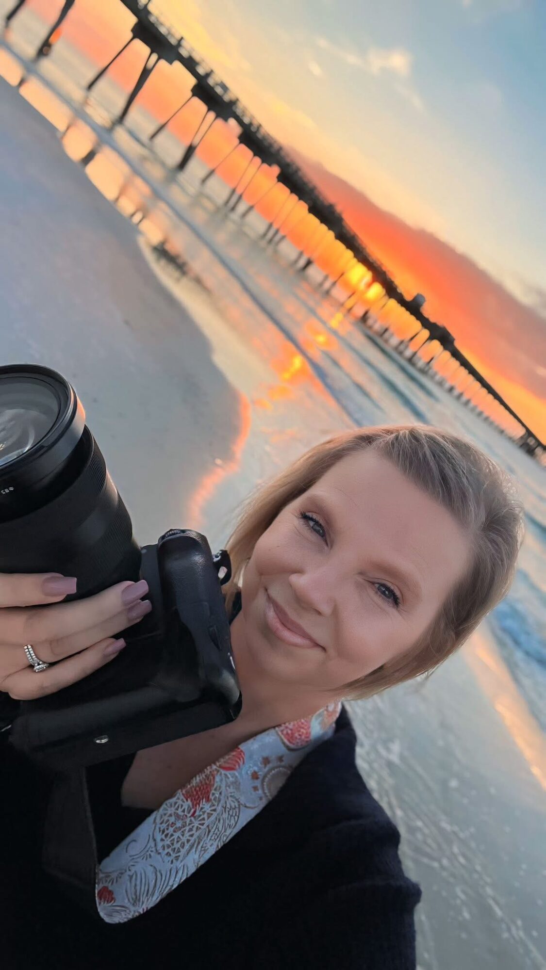 Woman taking a selfie near water with a bridge and sunset in the background.