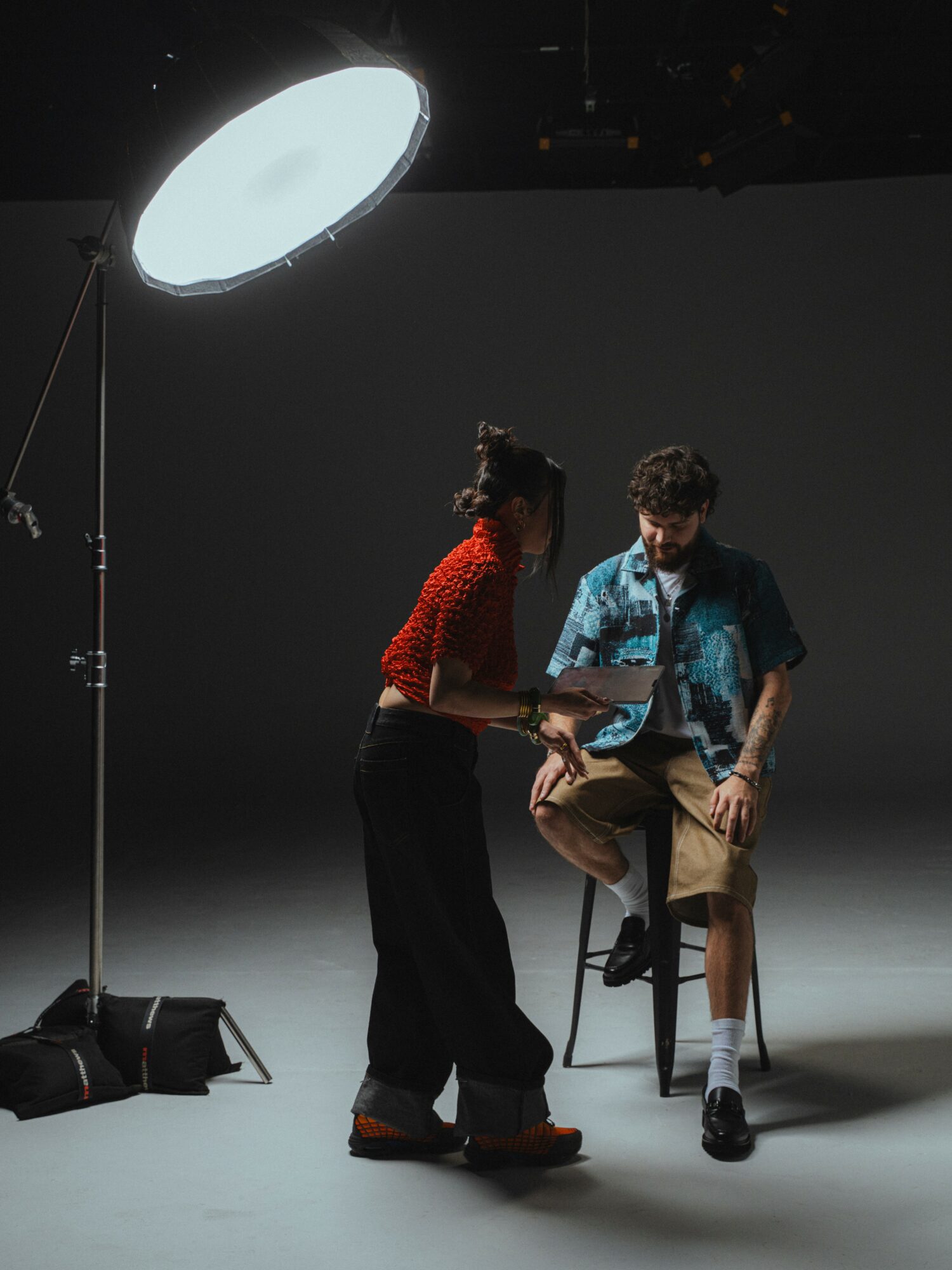 Two people interact on a dark stage with a large overhead light, one standing and one sitting on a stool, holding a tablet.