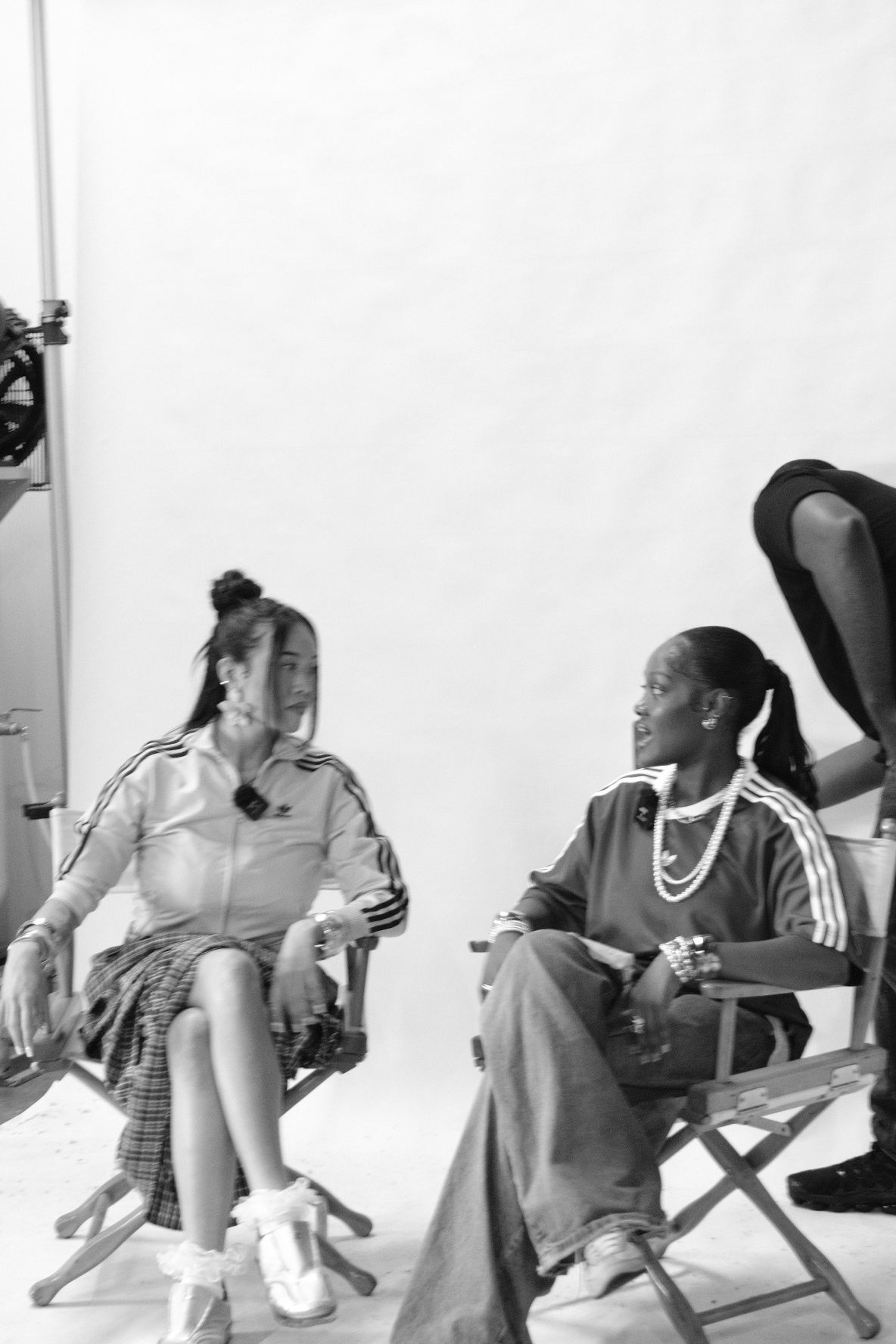 Two women sitting and standing in a room, one woman seated with a bag on her lap, the other standing nearby.