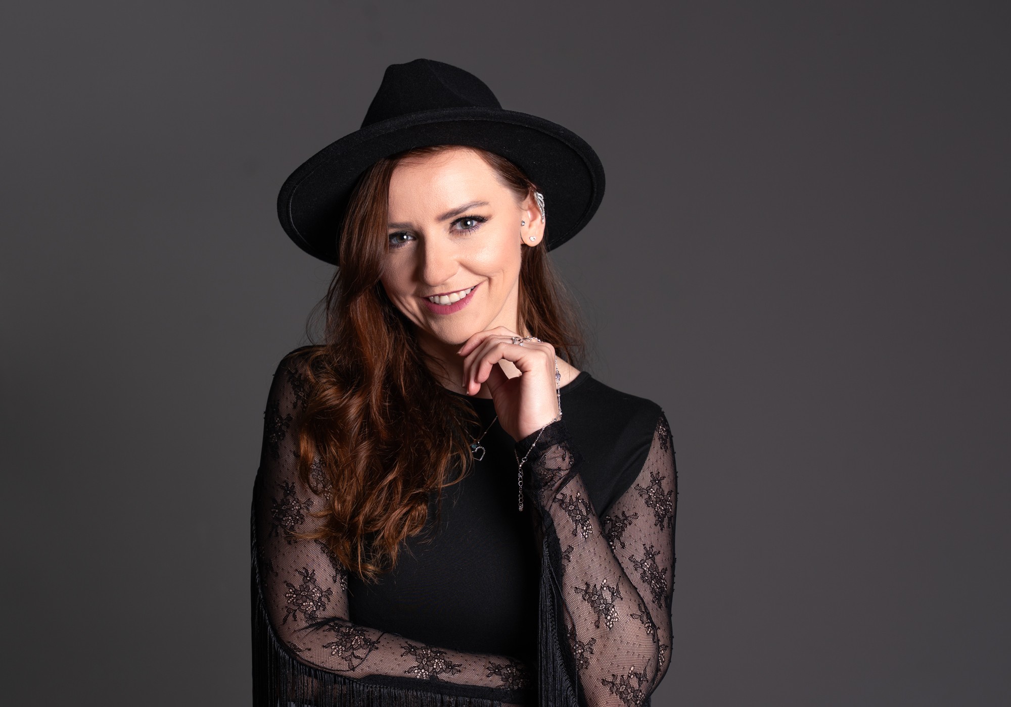 Woman with long hair wearing a black hat and lace top, smiling, against a gray background.