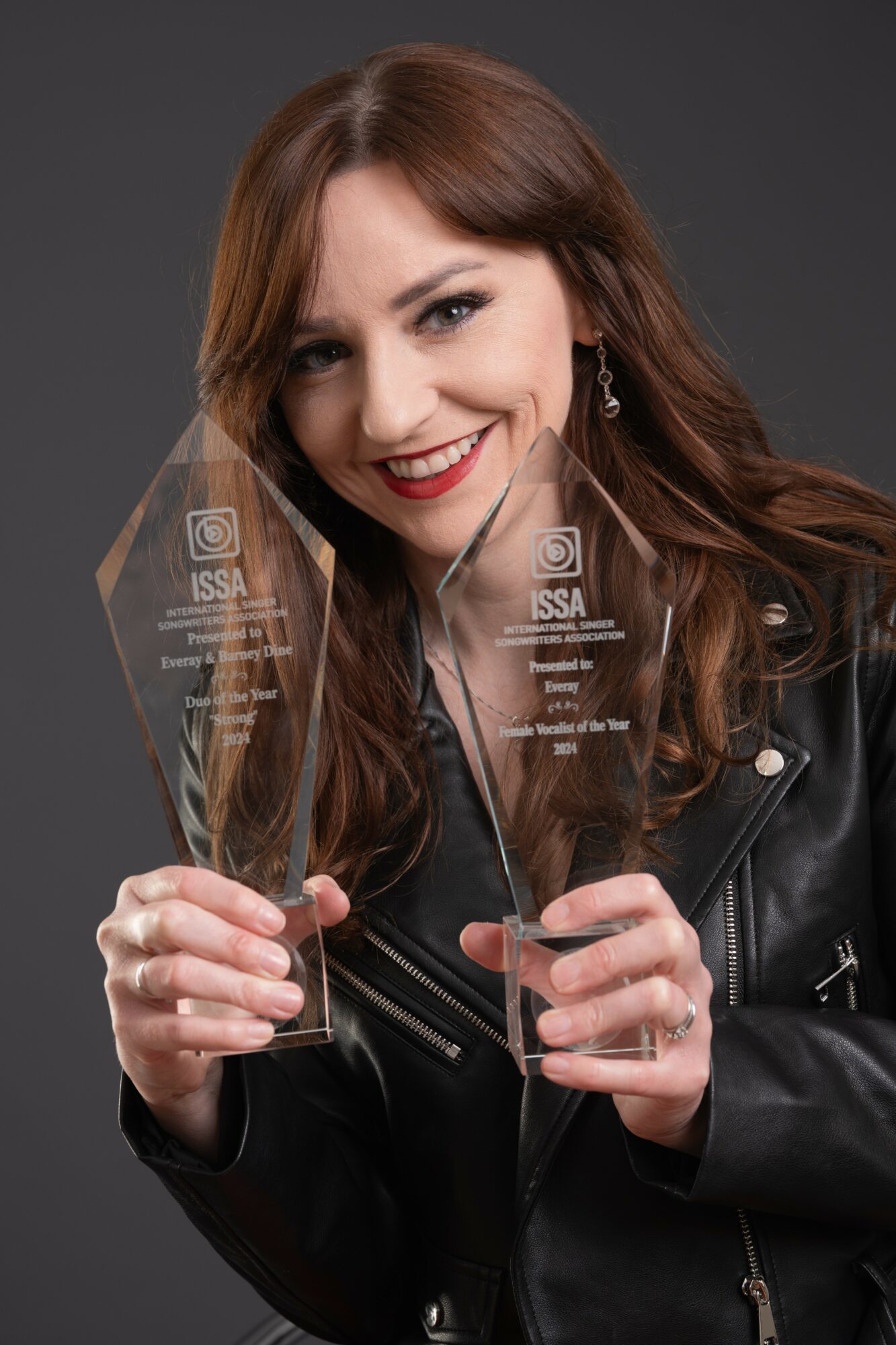 Woman with brown hair smiling, holding two clear awards, wearing a black leather jacket, against a dark background.