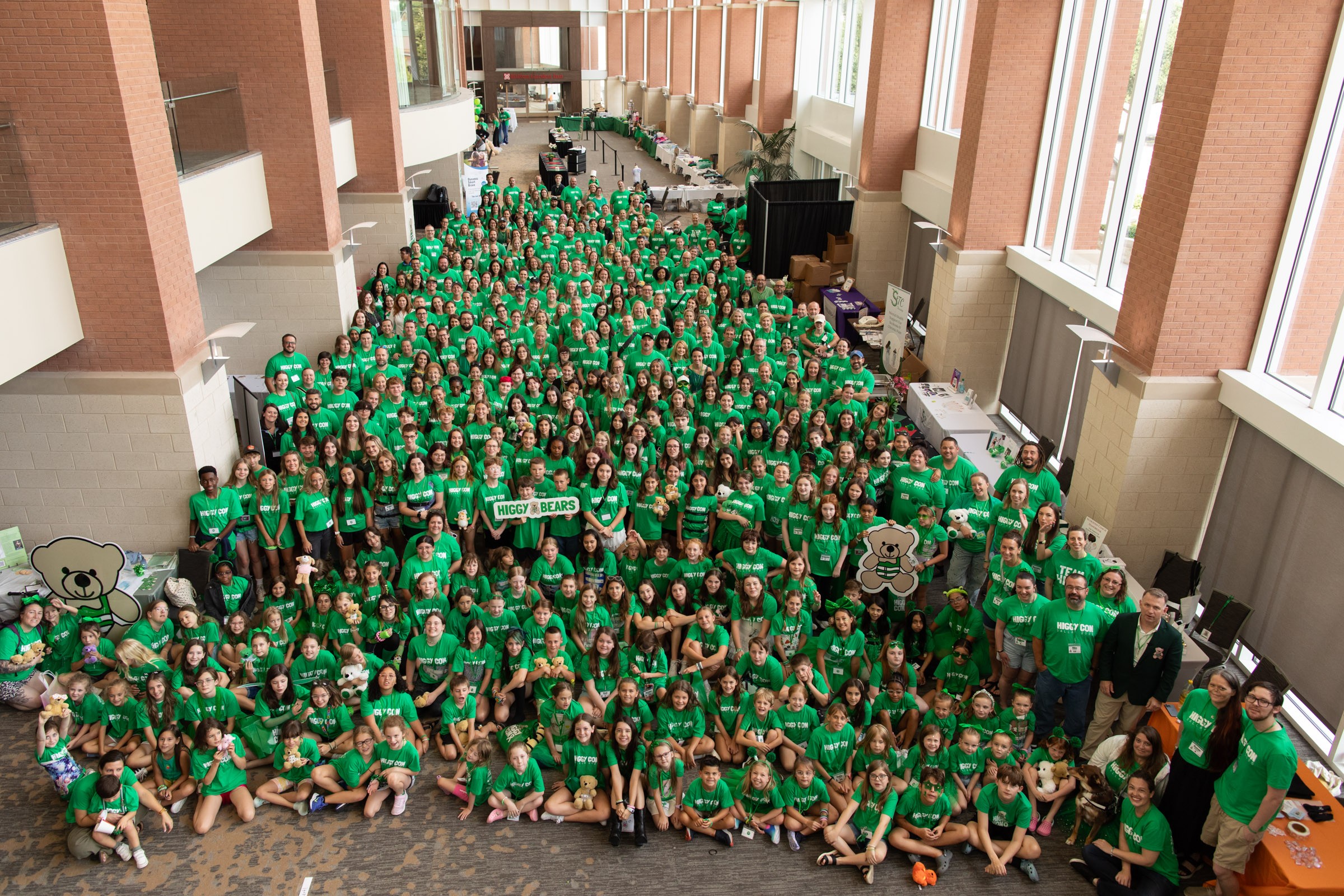 Large group of people wearing green shirts gathered in a spacious indoor area with tall windows and brick walls.