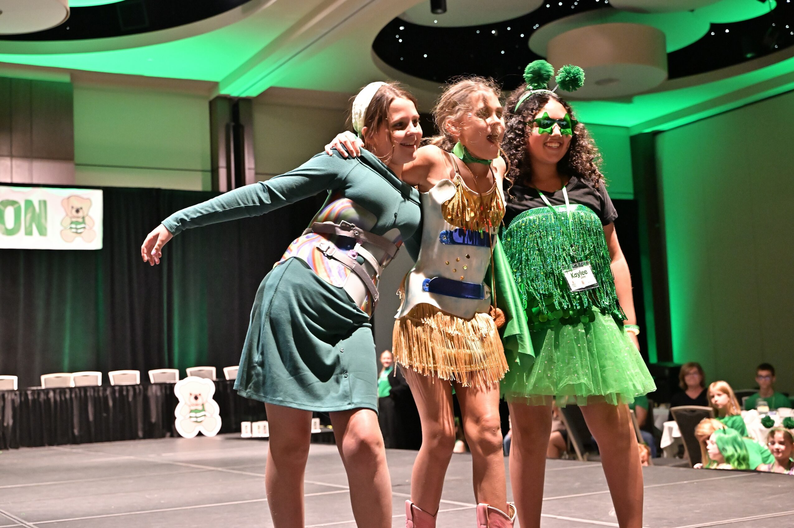 Four women in costumes pose on stage with arms around each other, smiling, in a green-lit event space.