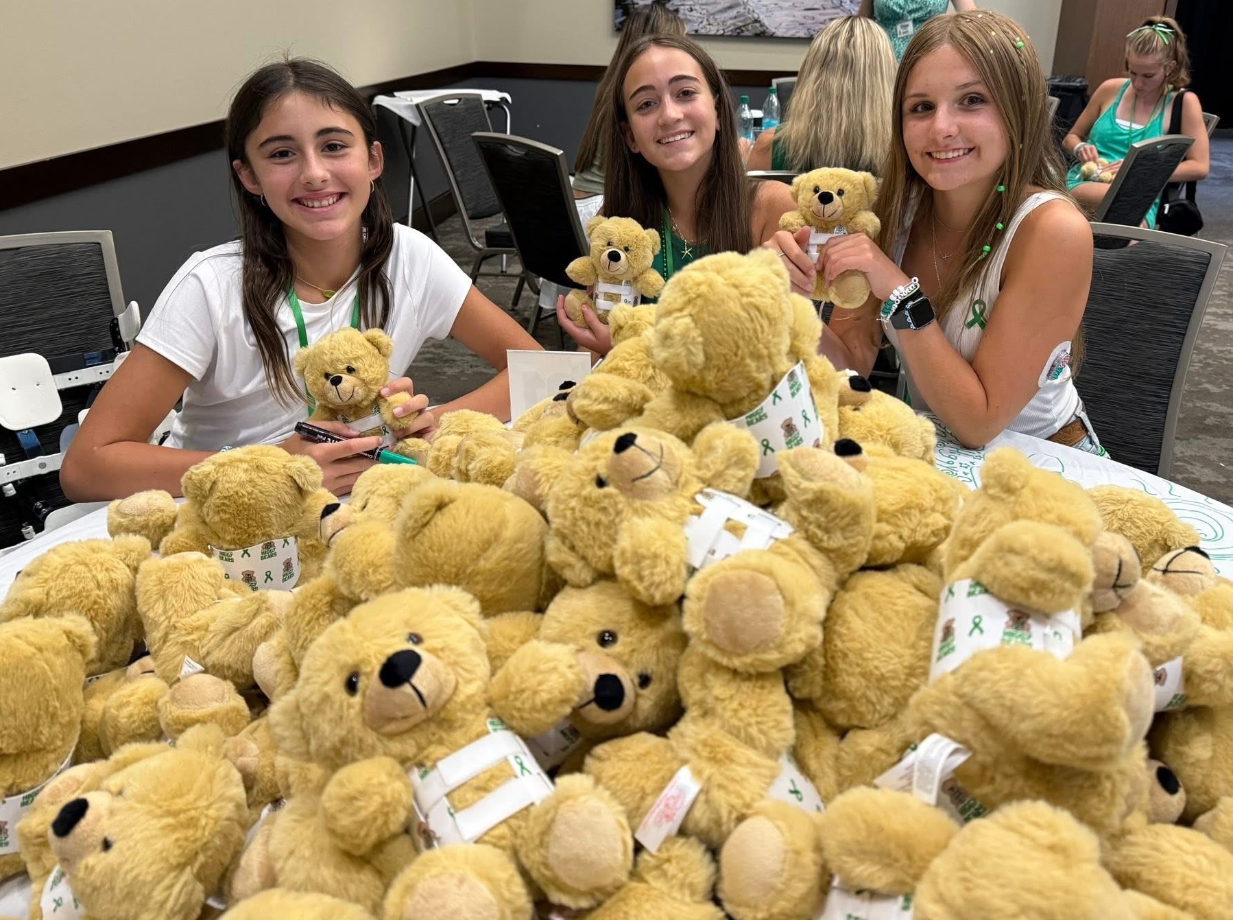 Three girls smiling behind a table filled with numerous teddy bears, some holding smaller bears, in a room with other people.