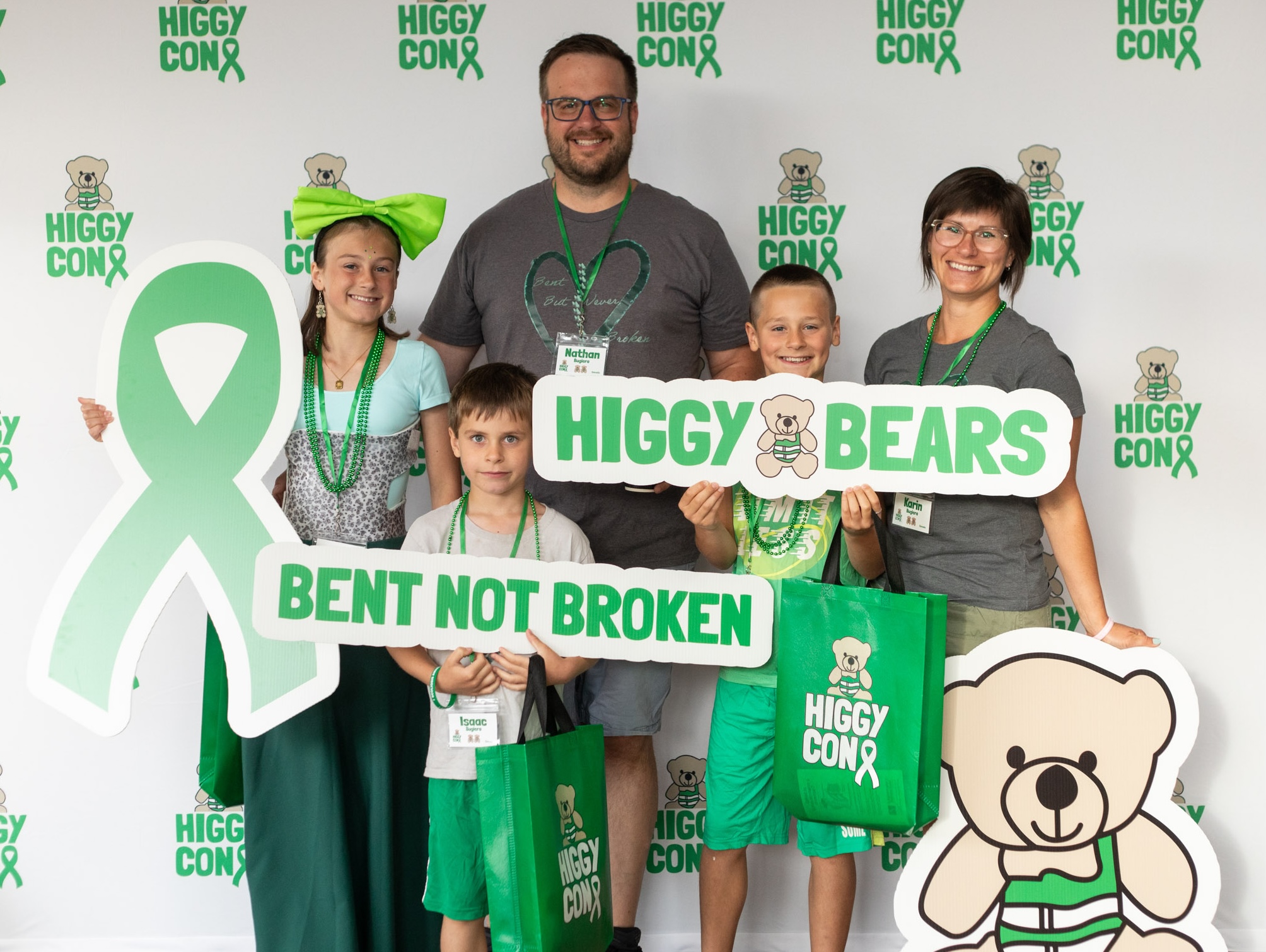 Group of five people holding signs and bags at Higgy Con, with a bear mascot and green ribbon background.
