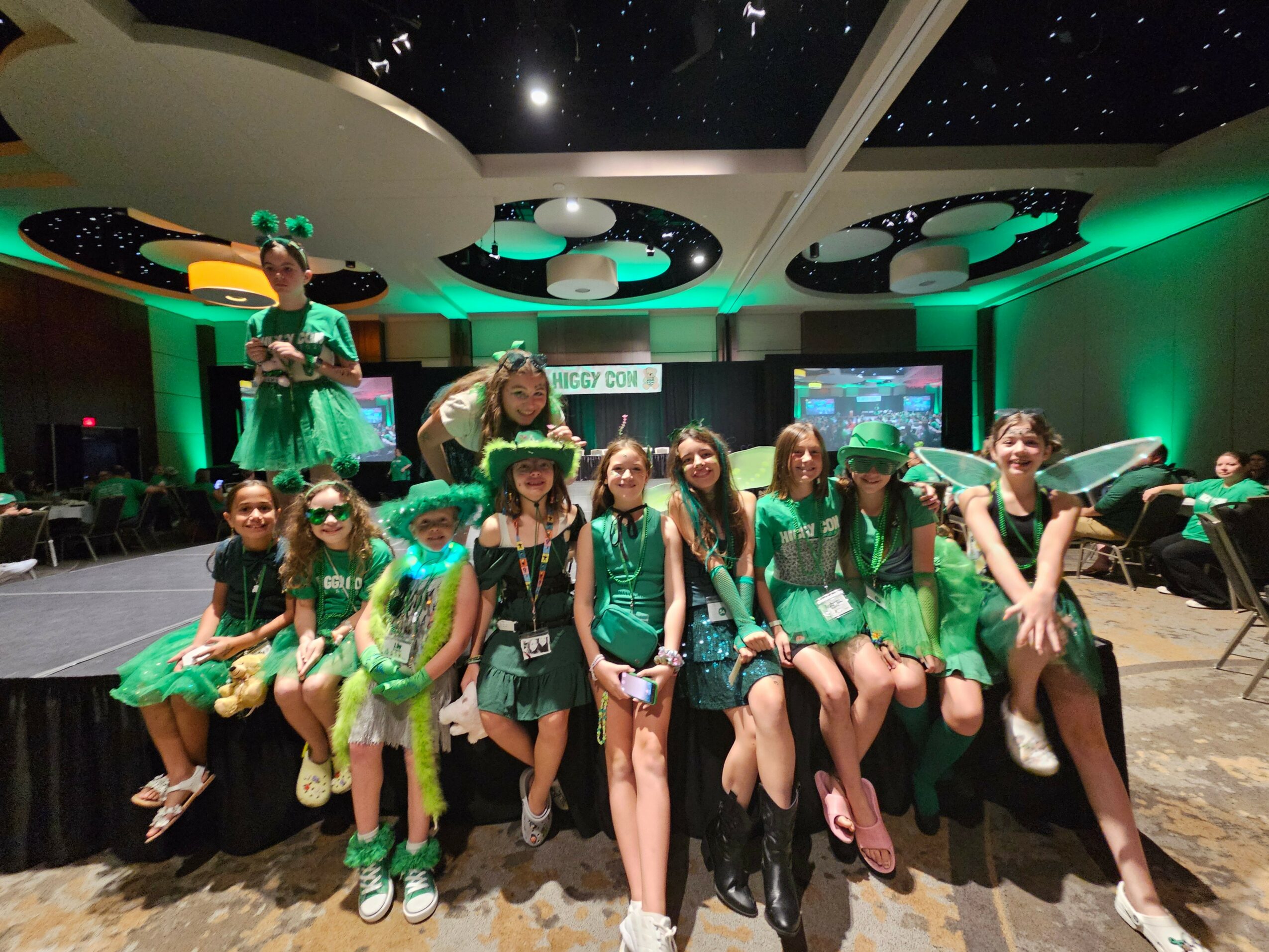 Group of children in green costumes sitting on a stage in a decorated room with a starry ceiling.